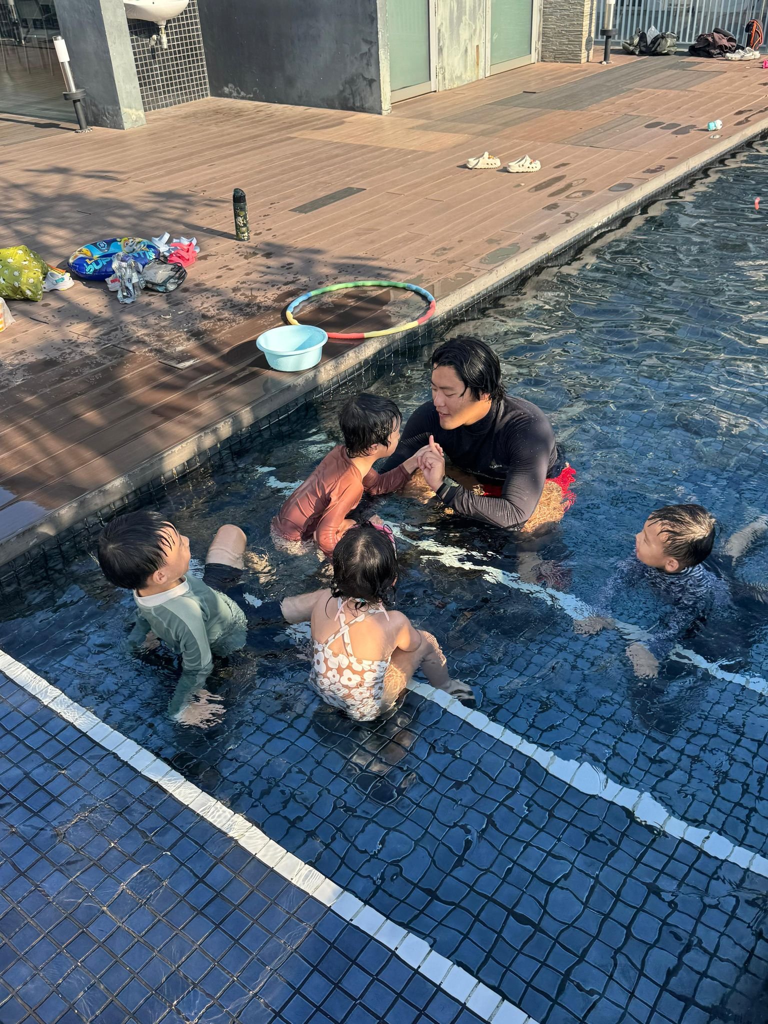 A man teaching four children how to swim in a swimming pool, with a wooden deck and some shoes and bags on the deck nearby.