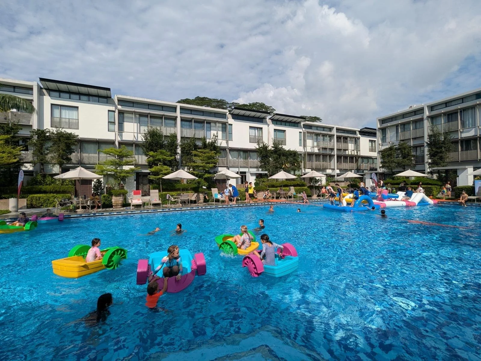 Children and adults swimming and playing in a large outdoor swimming pool with colorful floats, surrounded by a hotel or residential complex with lounge chairs and umbrellas.