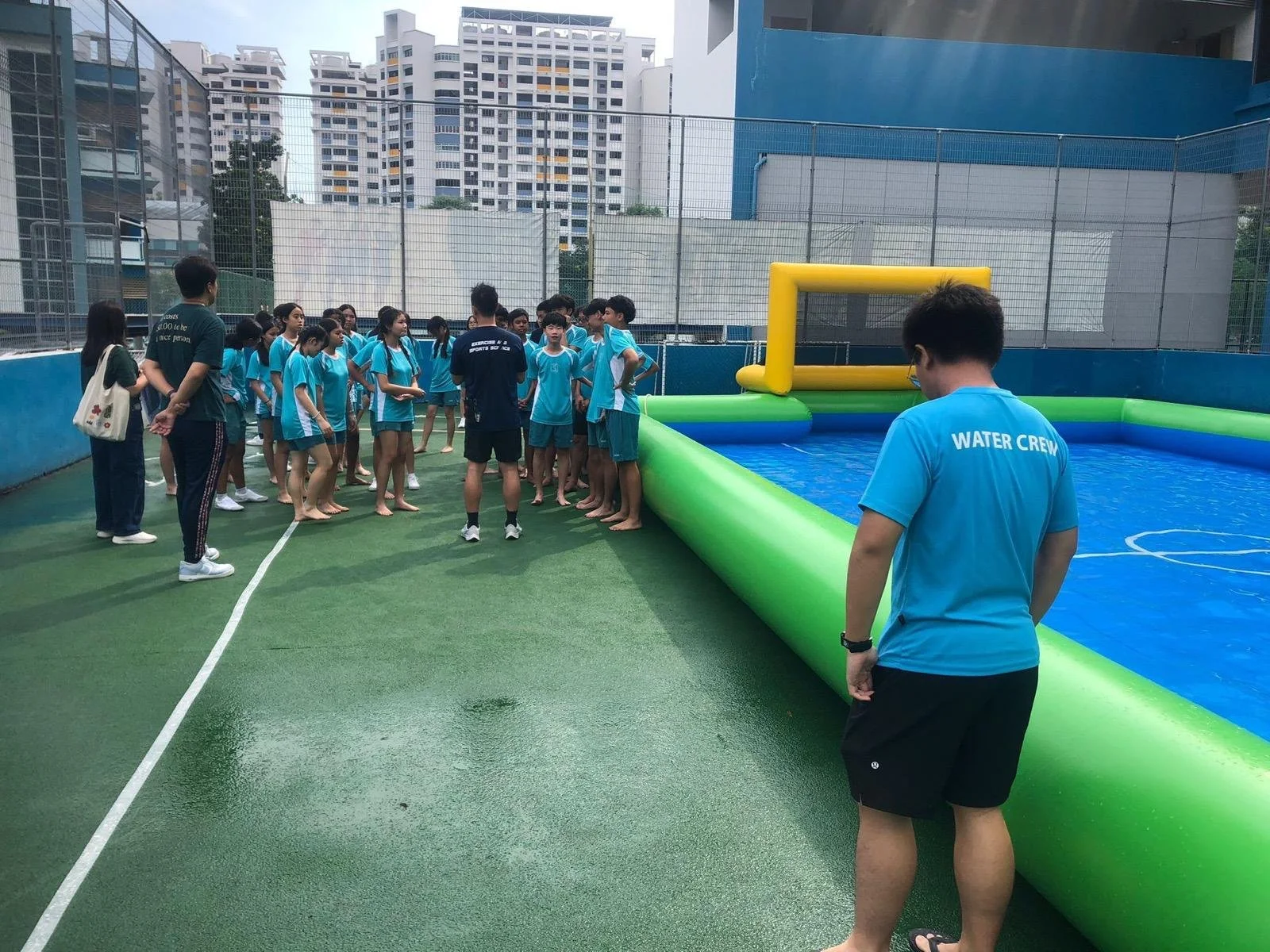 Group of children and adults gathered around a water activity pool on a rooftop, with a water crew member supervising, in an urban area with tall buildings in the background.