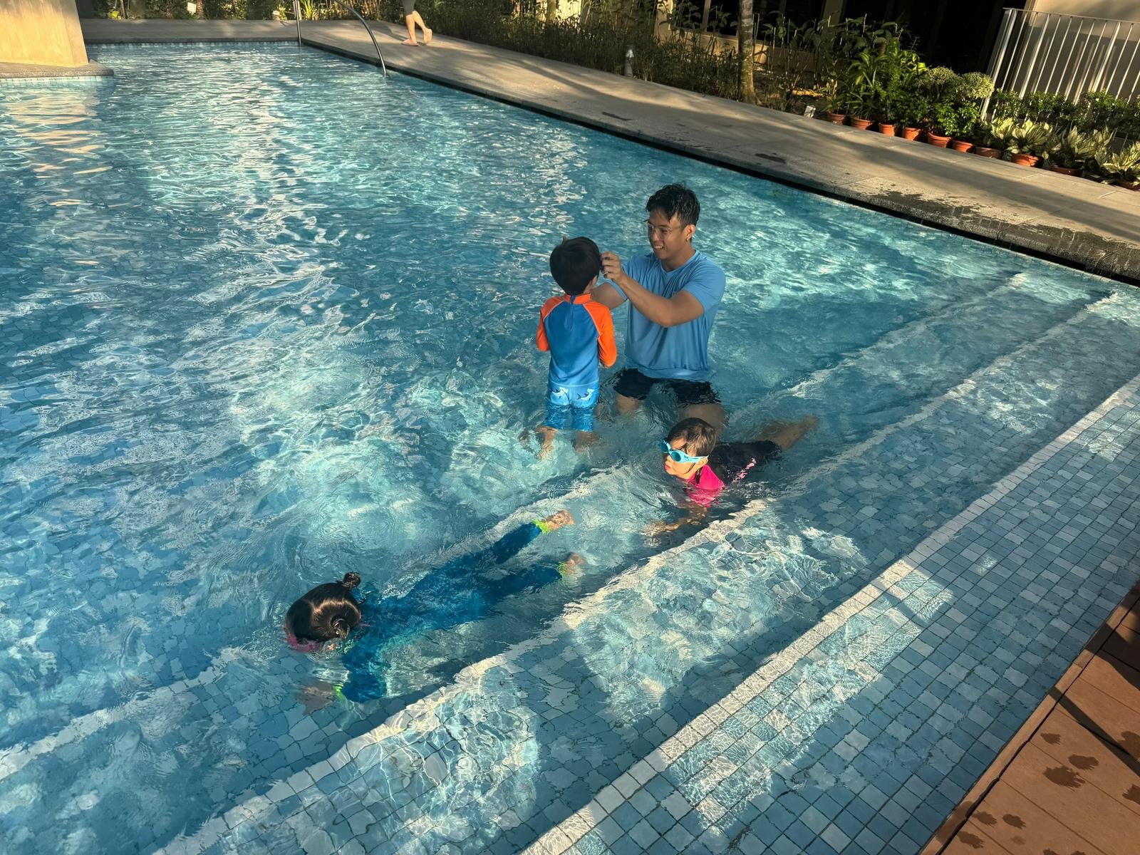 A man teaching two children how to swim in an outdoor pool on a sunny day. The children are wearing goggles and swimsuits, and the man is assisting a boy standing in the shallow end.