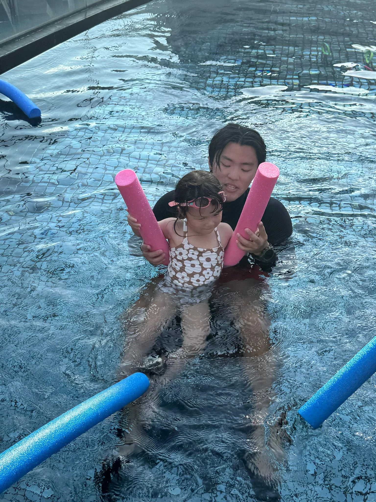 A man helping a young girl learn to swim in a pool, holding pink pool noodles for support.