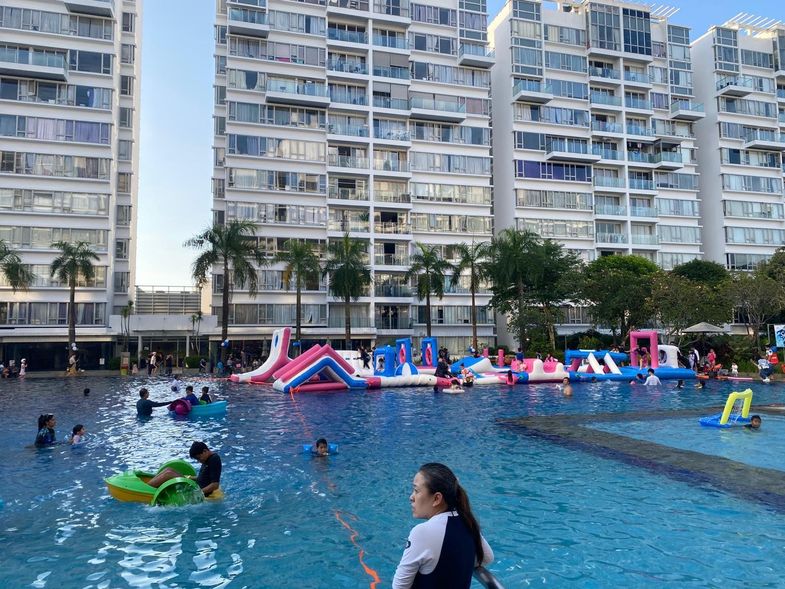 People swimming and playing in a large outdoor pool with inflatable slides and obstacles, surrounded by high-rise apartment buildings and palm trees.