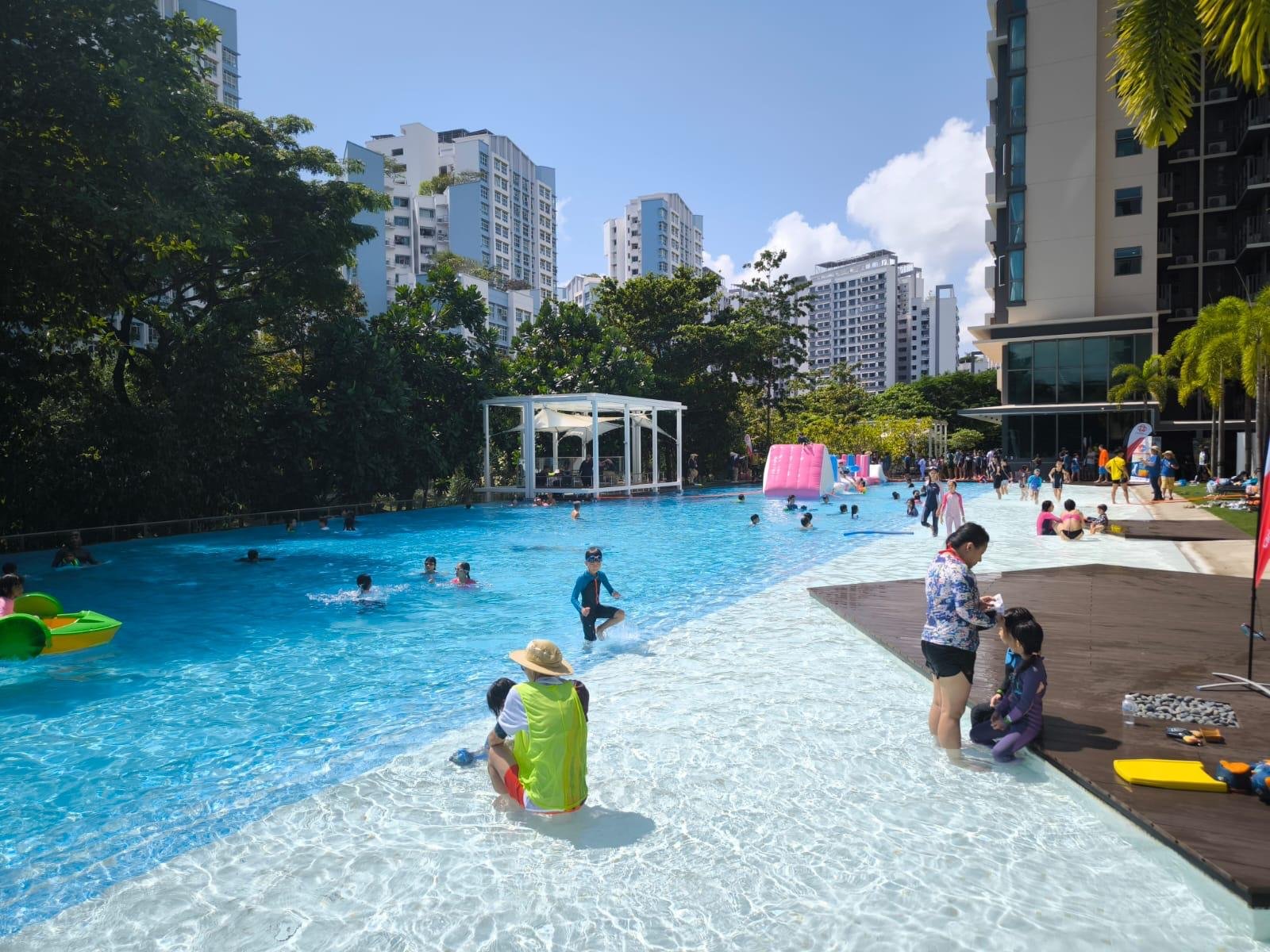 People swimming and playing at an outdoor pool surrounded by trees and tall buildings on a sunny day.