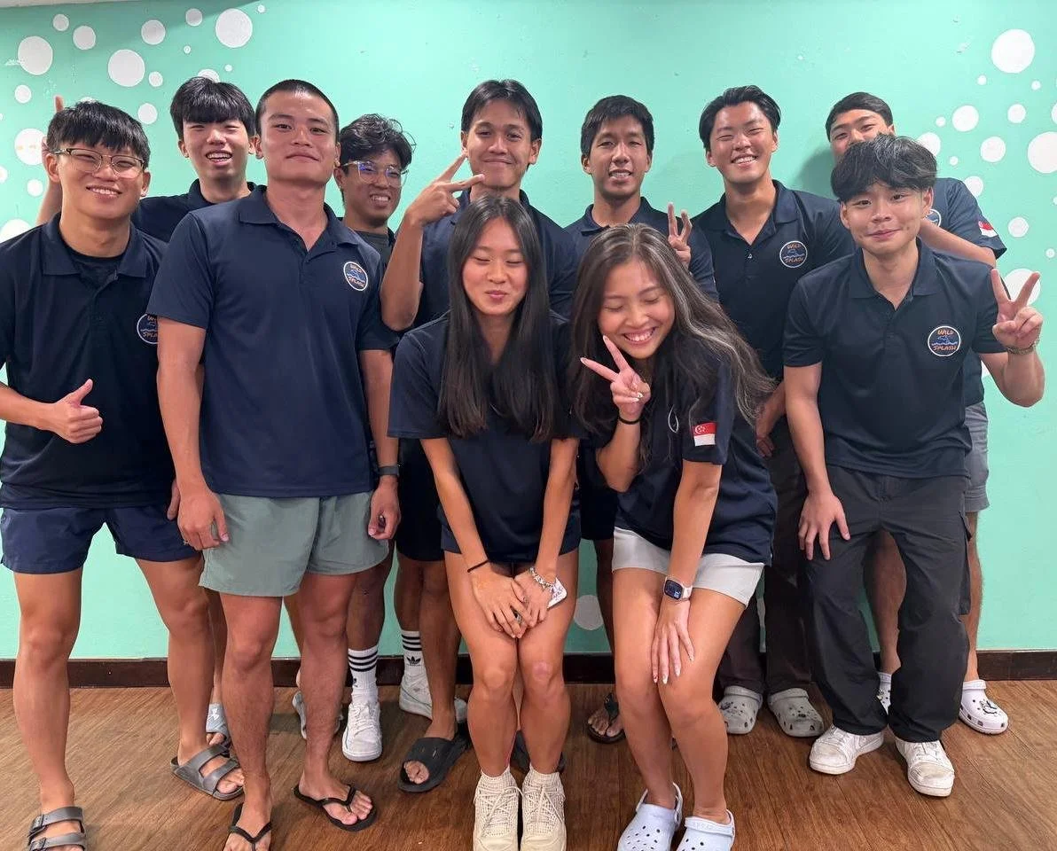 Group of twelve young people posing together indoors against a green wall with white circles, some showing peace signs and others smiling.