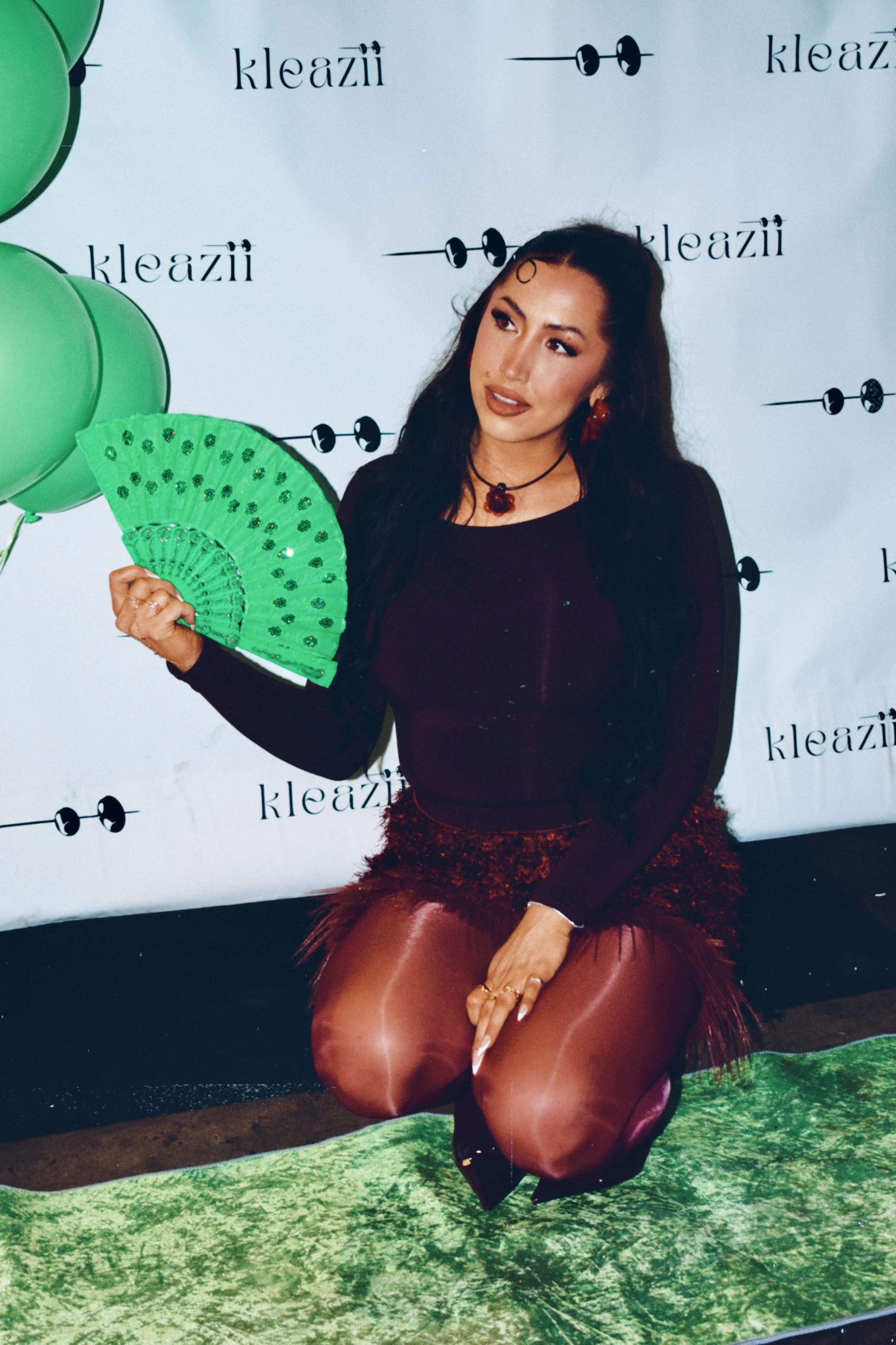 Woman wearing dark top and leather pants, holding a green decorative fan, kneeling on a green surface, with a backdrop featuring black text and graphics, and green balloons on the left.