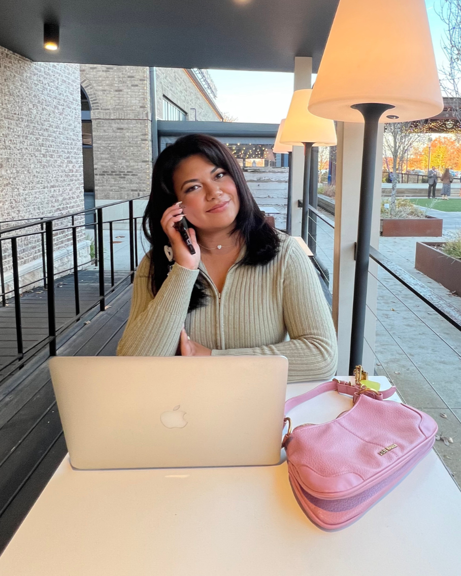 A woman with dark hair, wearing a sweater, sitting at a white table in an outdoor cafe, talking on her cell phone. On the table are a silver MacBook and a pink purse. The scene is set in an urban area with a brick building, outdoors with trees and a few people in the background, during autumn.