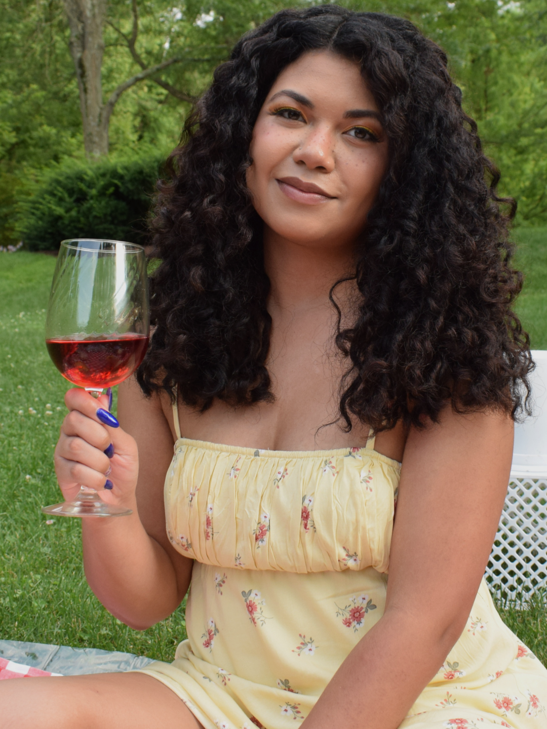 Young woman with curly dark hair holding a glass of red wine outdoors in a garden.
