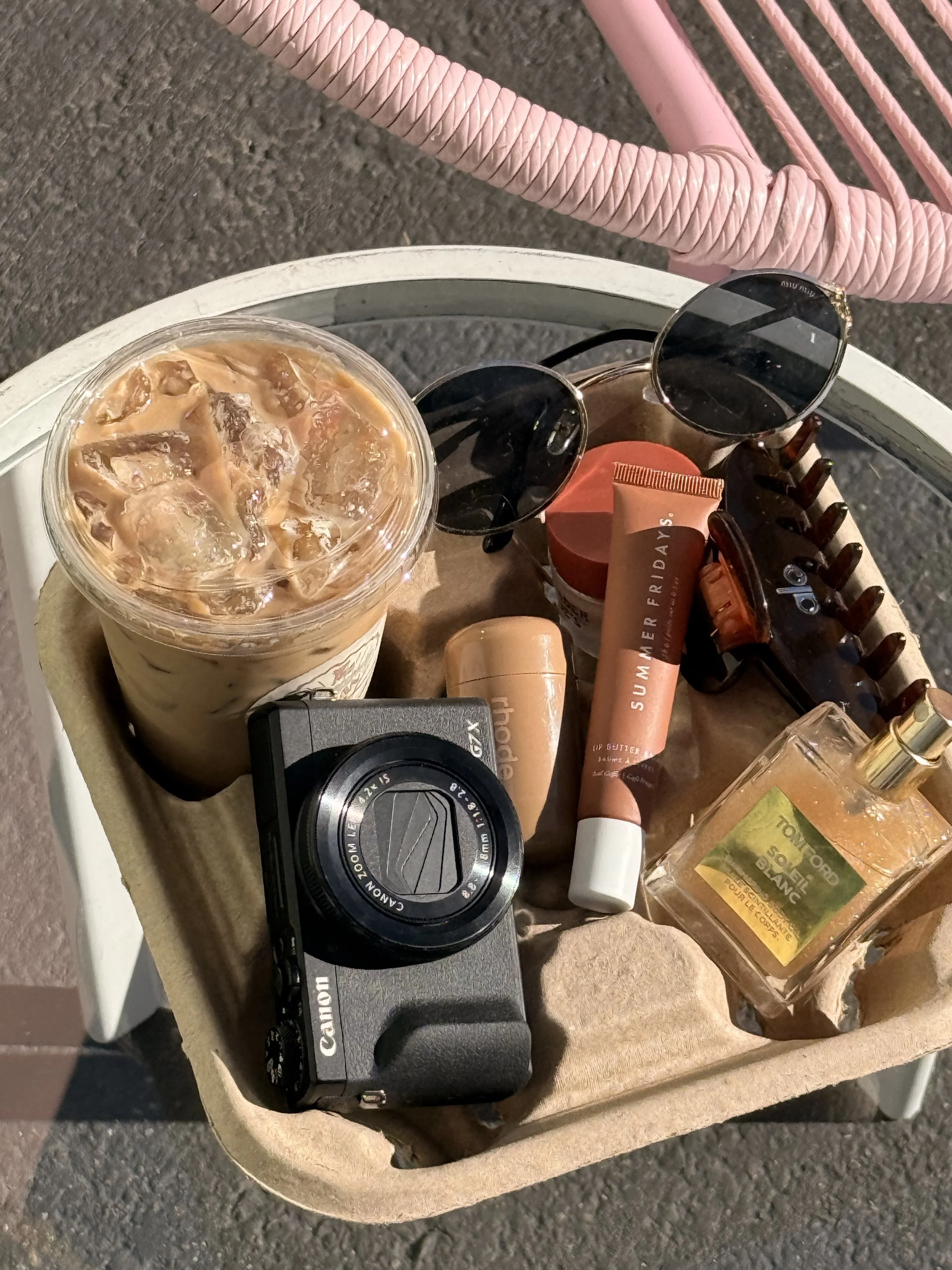 A round beige sidewalk table with various items on it, including a large iced coffee, a pair of round sunglasses, a small jar of moisturizer, a tube of sunscreen, a hairbrush, a bottle of perfume, and a camera lens cap, with a pink woven chair and gray pavement in the background.