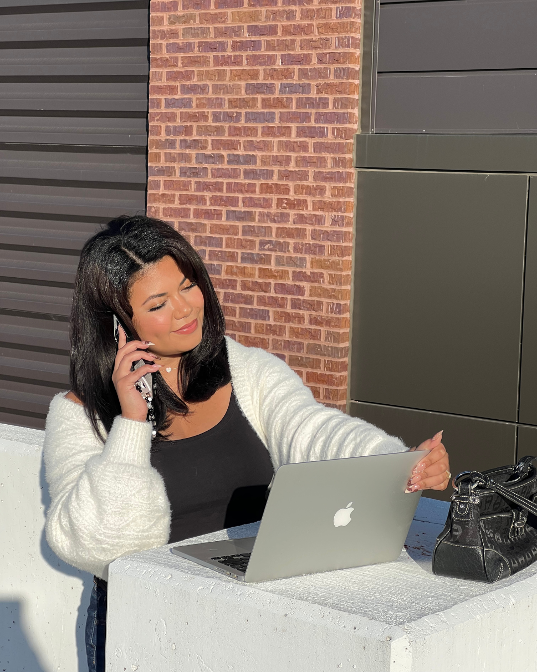A woman with dark hair, wearing a white sweater and black top, sitting at a white table outdoors. She is talking on a cell phone and looking at a silver MacBook. A black purse is on the table.