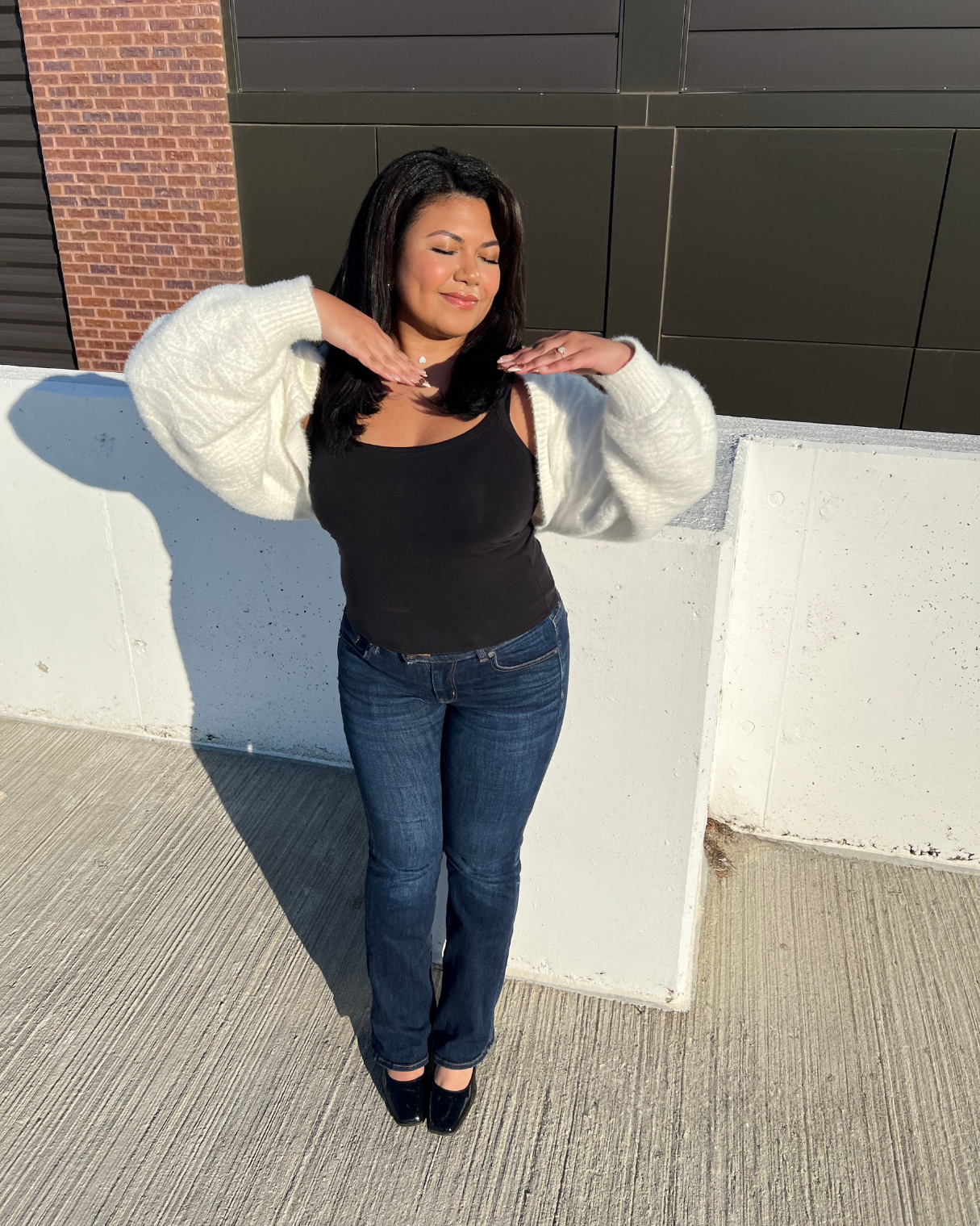 Woman with dark hair, wearing a black top, blue jeans, and a white cardigan, standing outdoors with eyes closed and hands near her shoulders, sunlight casting a shadow on a white concrete wall behind her.