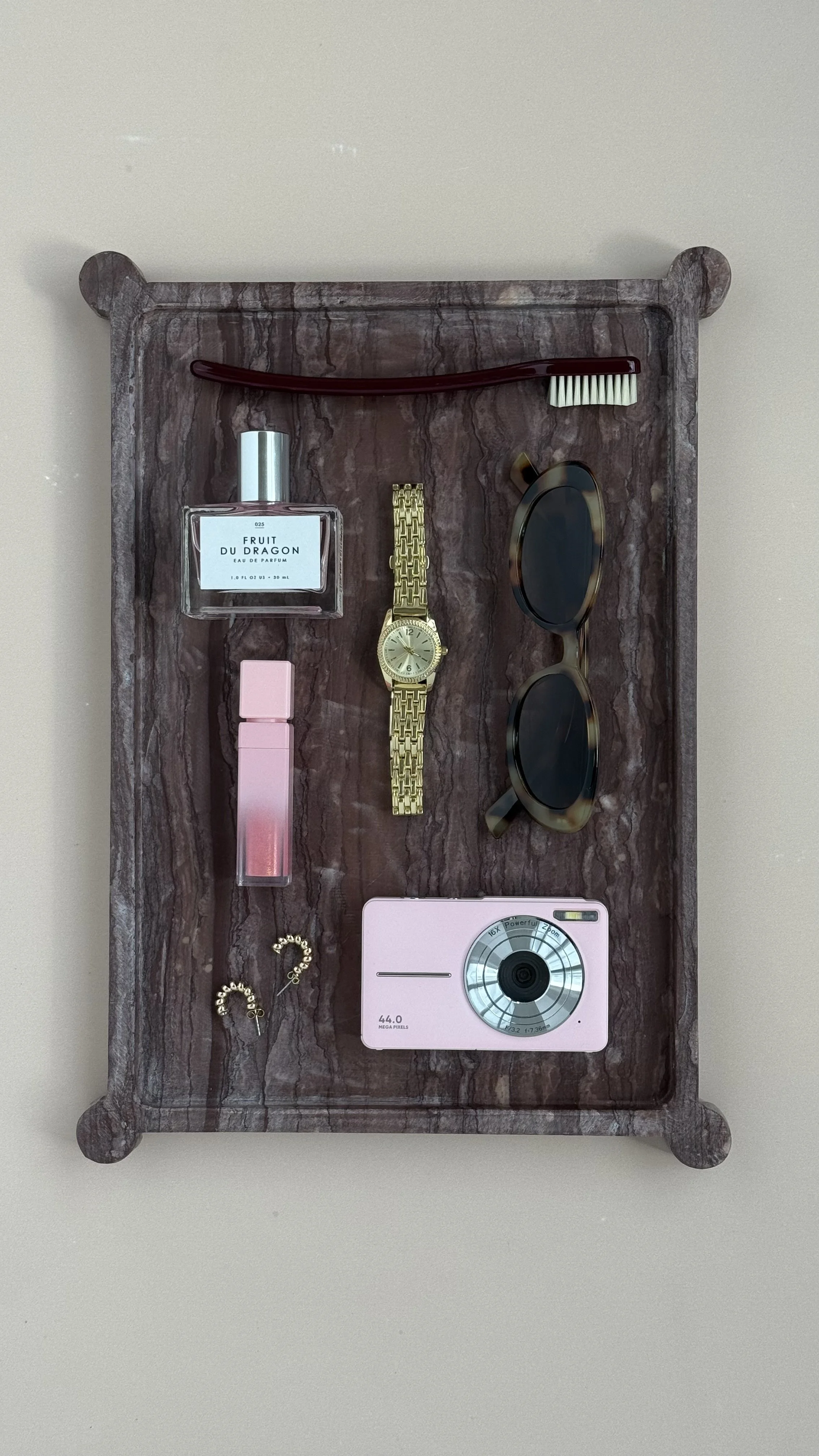 A marble tray holding a perfume bottle labeled 'Fruit Du Dragon,' a gold wristwatch, a pair of sunglasses, a pink lipstick, a small pink box, a makeup brush with a red handle, and a pink digital camera.