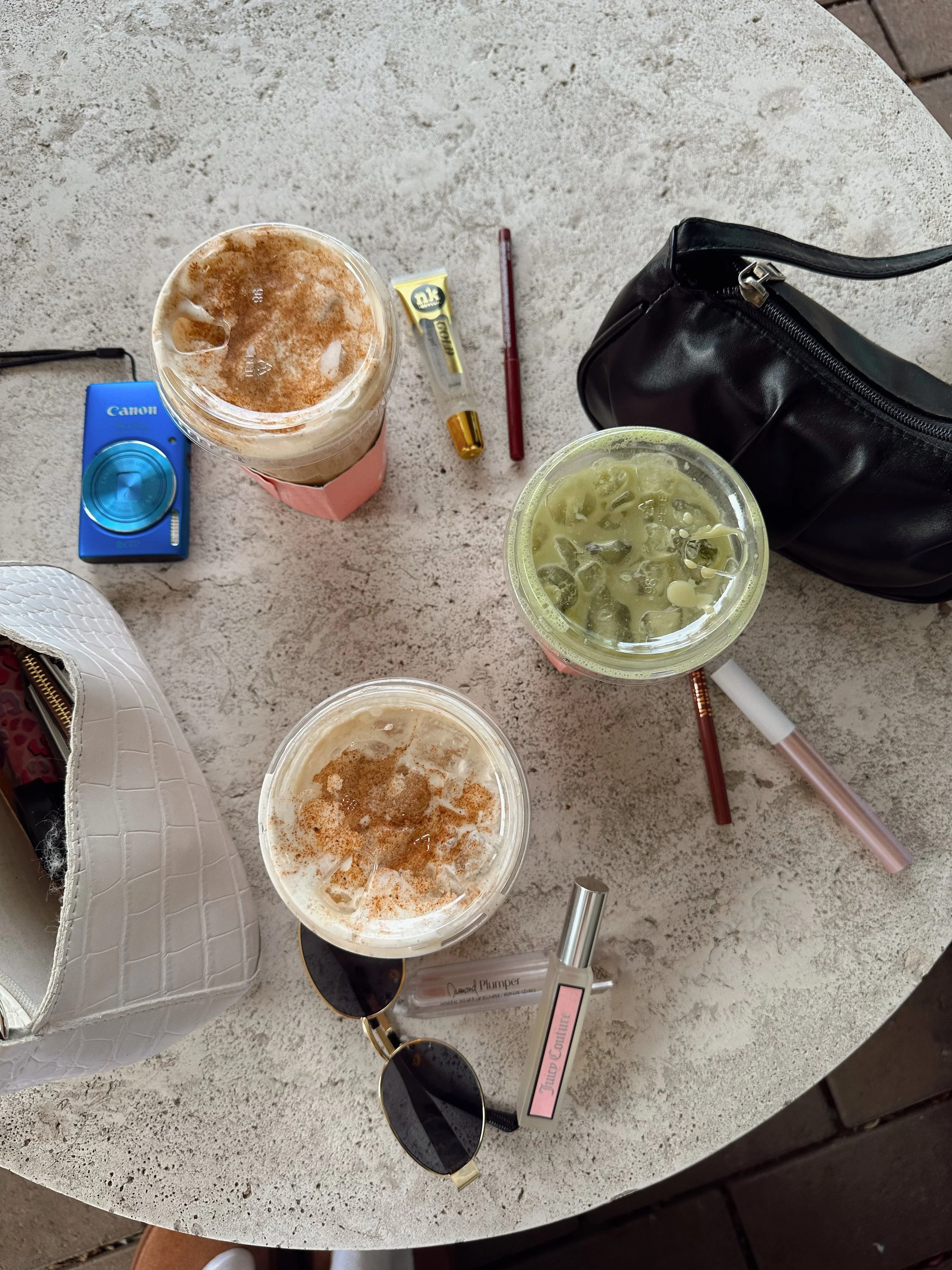 Table with two iced coffee drinks, sunglasses, a tube of lip balm, a black handbag, a white purse with a crocodile pattern, and a few pens.