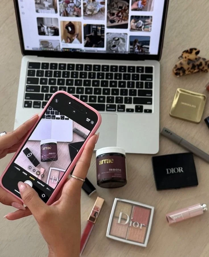 A person taking a photo of makeup products on a table with a smartphone. The table also has a MacBook displaying a gallery of images, a shell-shaped accessory, a gold case, a gray pen, and some other makeup items including a Dior palette and pink lipstick.