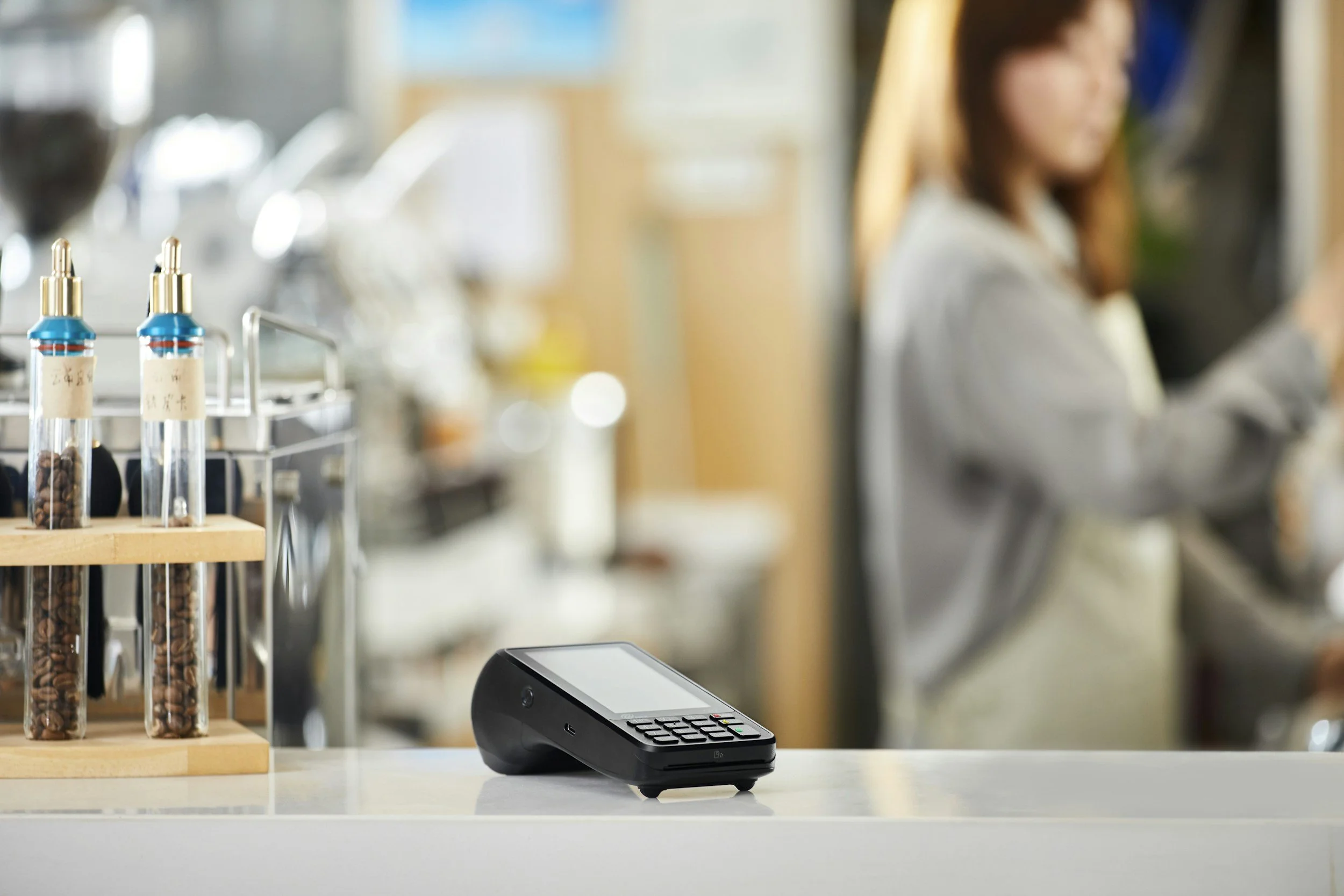A close-up shot of an Eftpos payment terminal sitting on a countertop, with a retail worker softly blurred in the background, representing digital payment infrastructure and merchant solutions.