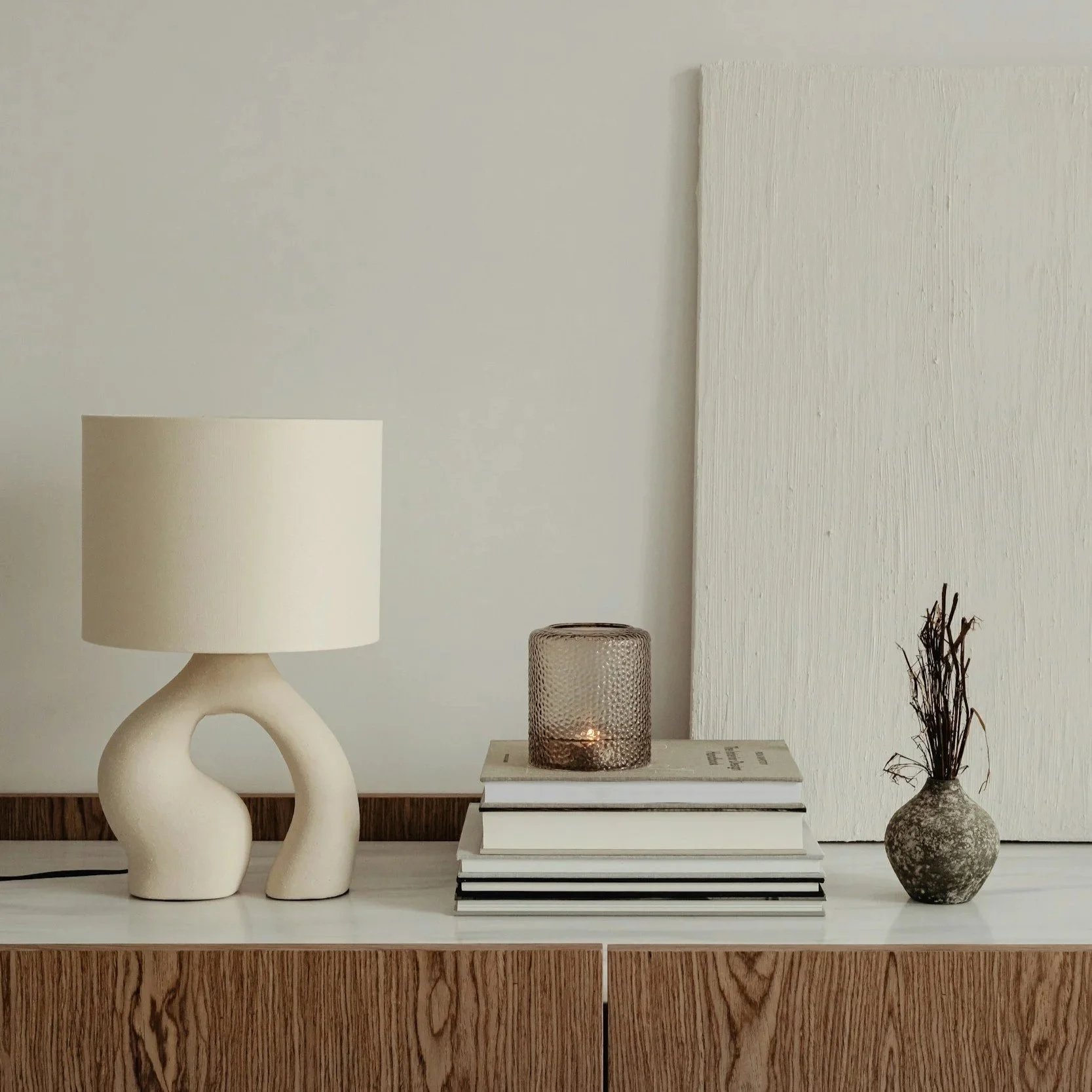 Koriste-esineitä pöydällä: valkoinen lampi, kokoelma kirjoja ja kynttilä, sekä savinen maljakko kuivuneilla oksilla