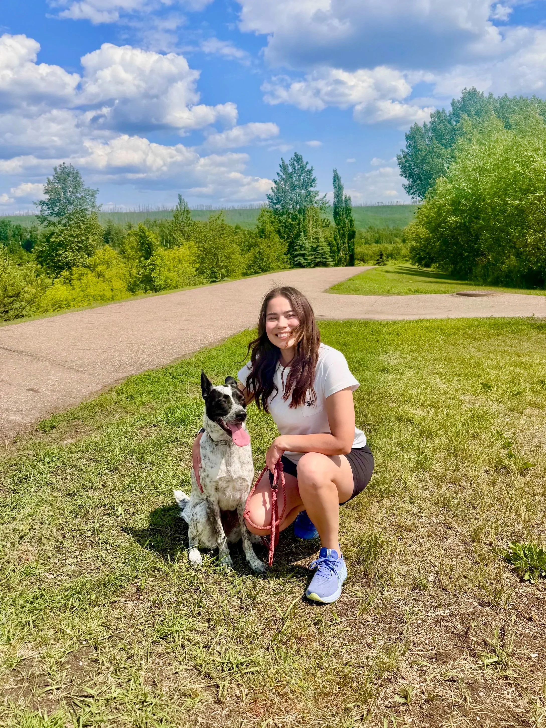 A woman in athletic clothing kneeling on grass next to a spotted black and white dog in a park with green trees and a blue sky with clouds.