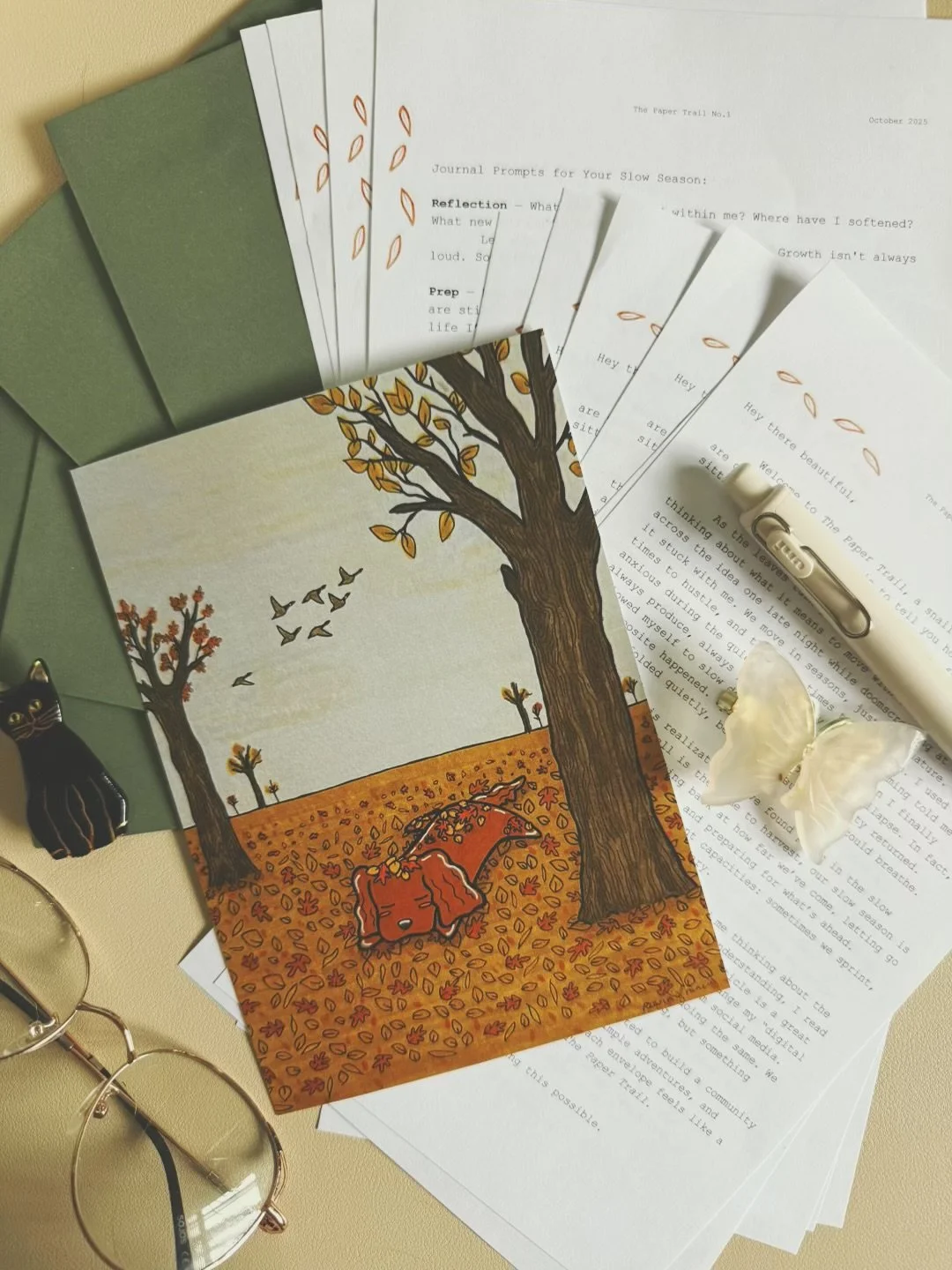 A colorful illustration of a fall scene with two trees and a dog laying on the ground, surrounded by fallen leaves. The scene is set on top of several sheets of paper with printed text, a white butterfly-shaped paper decoration, a white pen, a black cat figurine, glasses, and some envelopes.