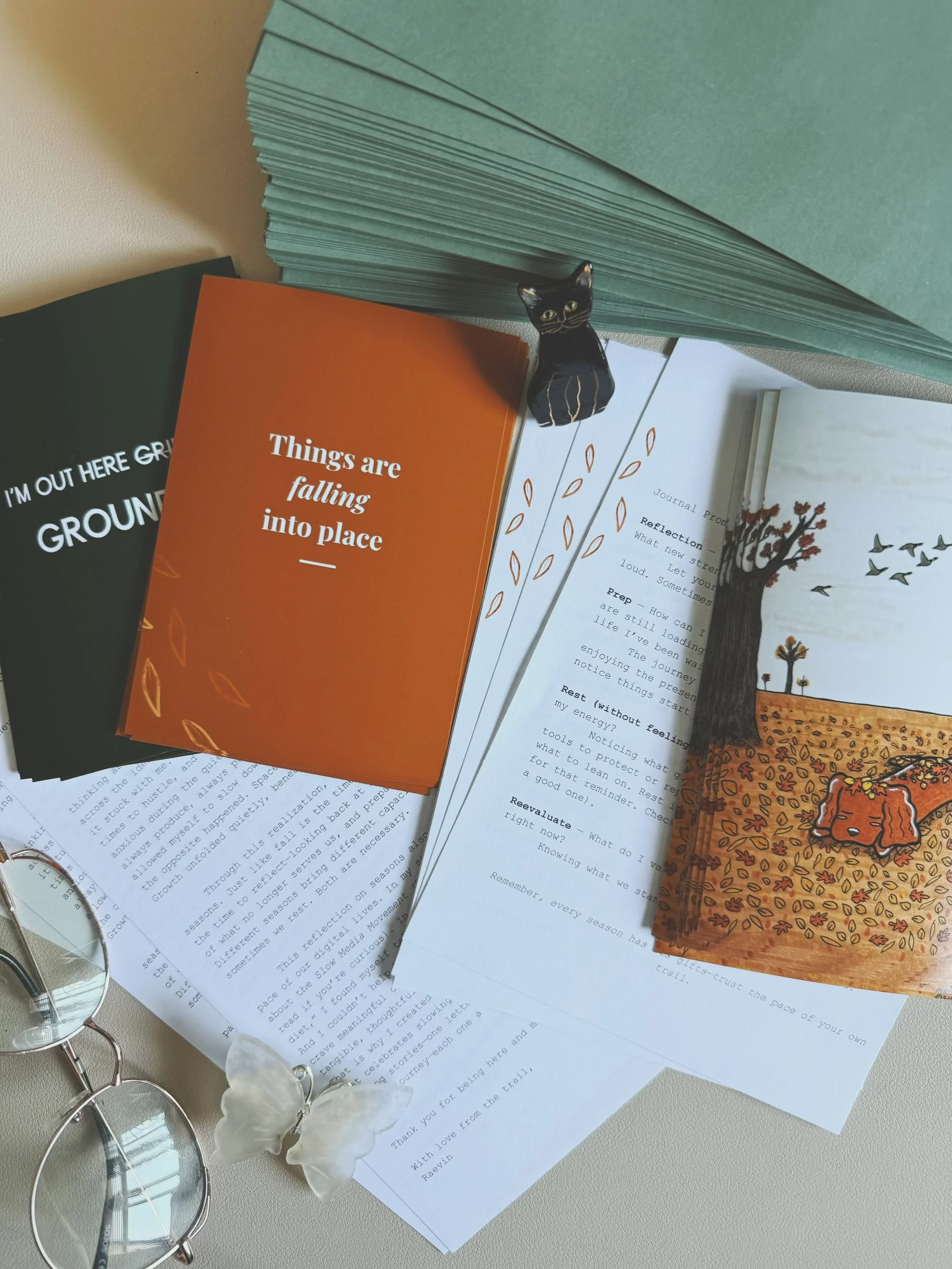Arrangement of colored paper, an illustrated book, a black cat figurine, papers with text, a pair of glasses, and a butterfly clip on a beige surface.
