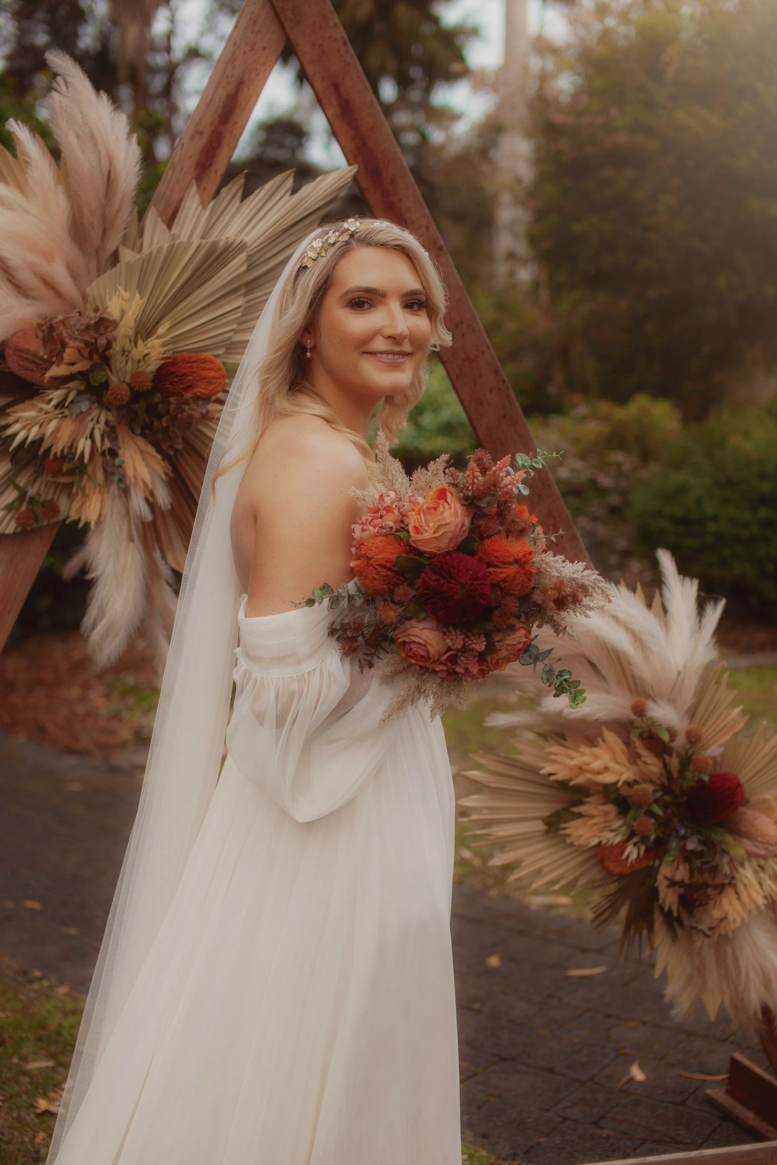 A bride in a white wedding dress holding a bouquet of orange and red flowers, standing outdoors with trees in the background, near a wooden floral display.