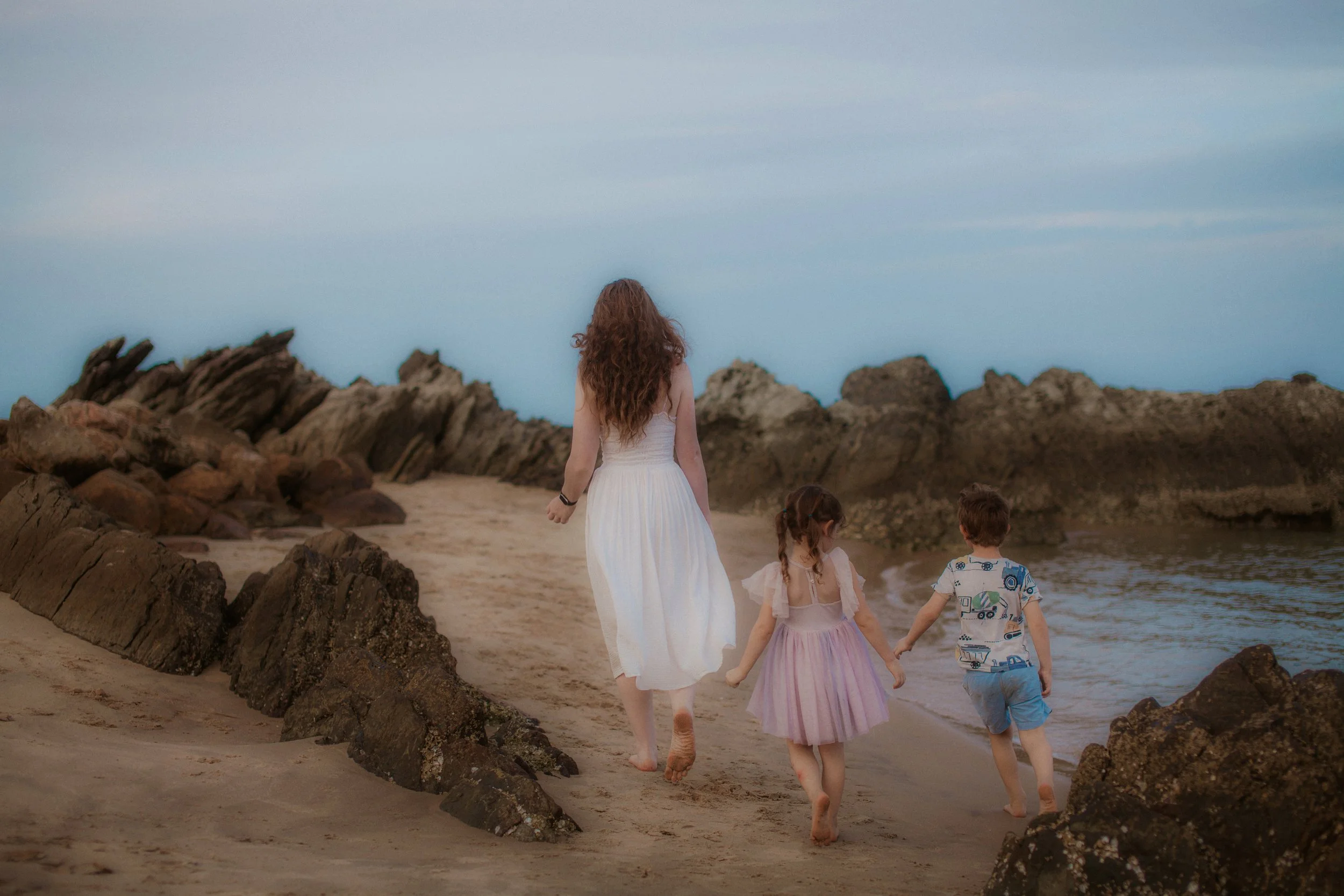 A woman and two children walking barefoot on a sandy beach near rocks, facing the ocean, with a cloudy sky overhead.