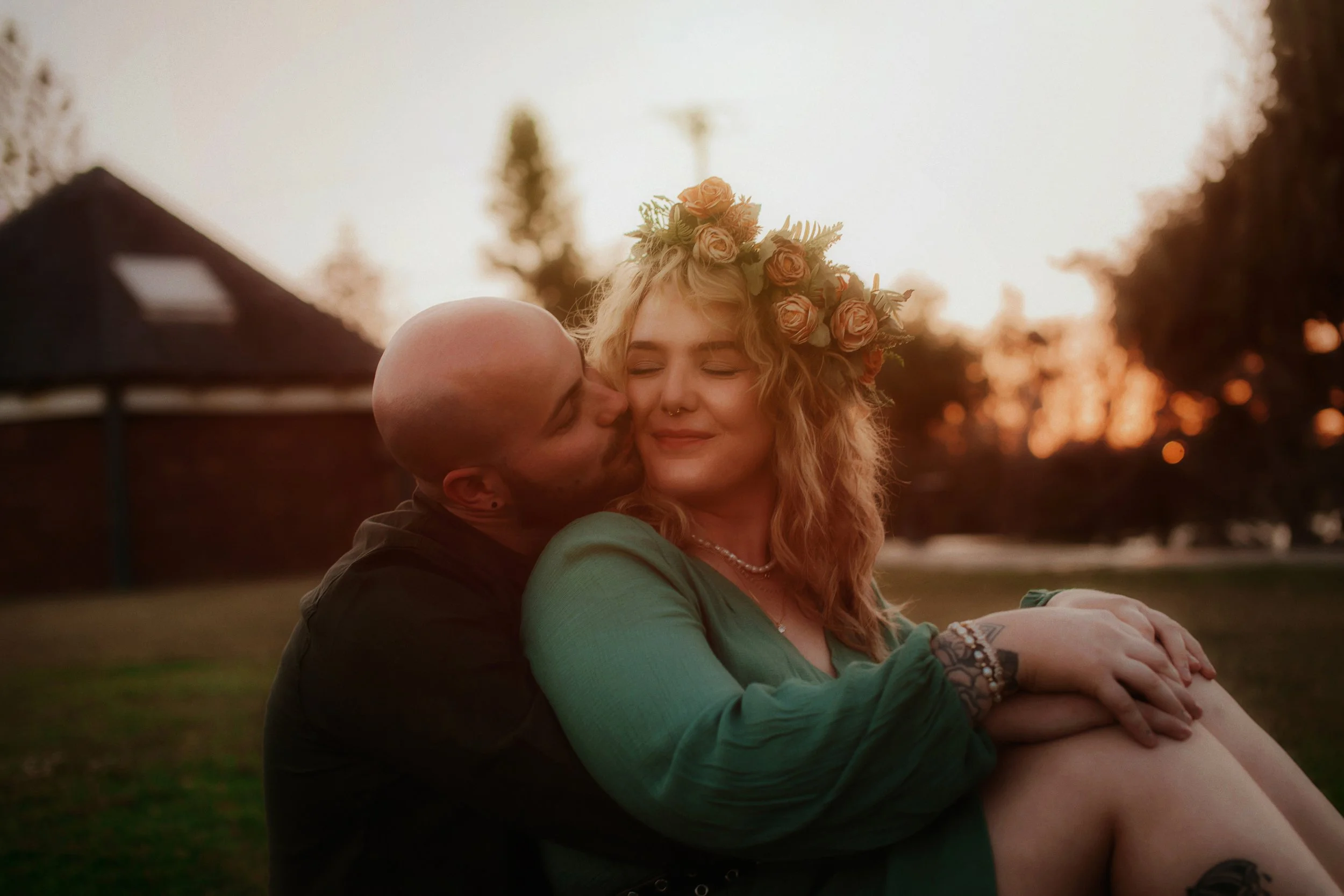 A couple sitting outdoors at sunset, with the man kissing the woman on the cheek. The woman has curly hair, a flower crown, and a content smile. They are embracing, with trees and a building in the background.