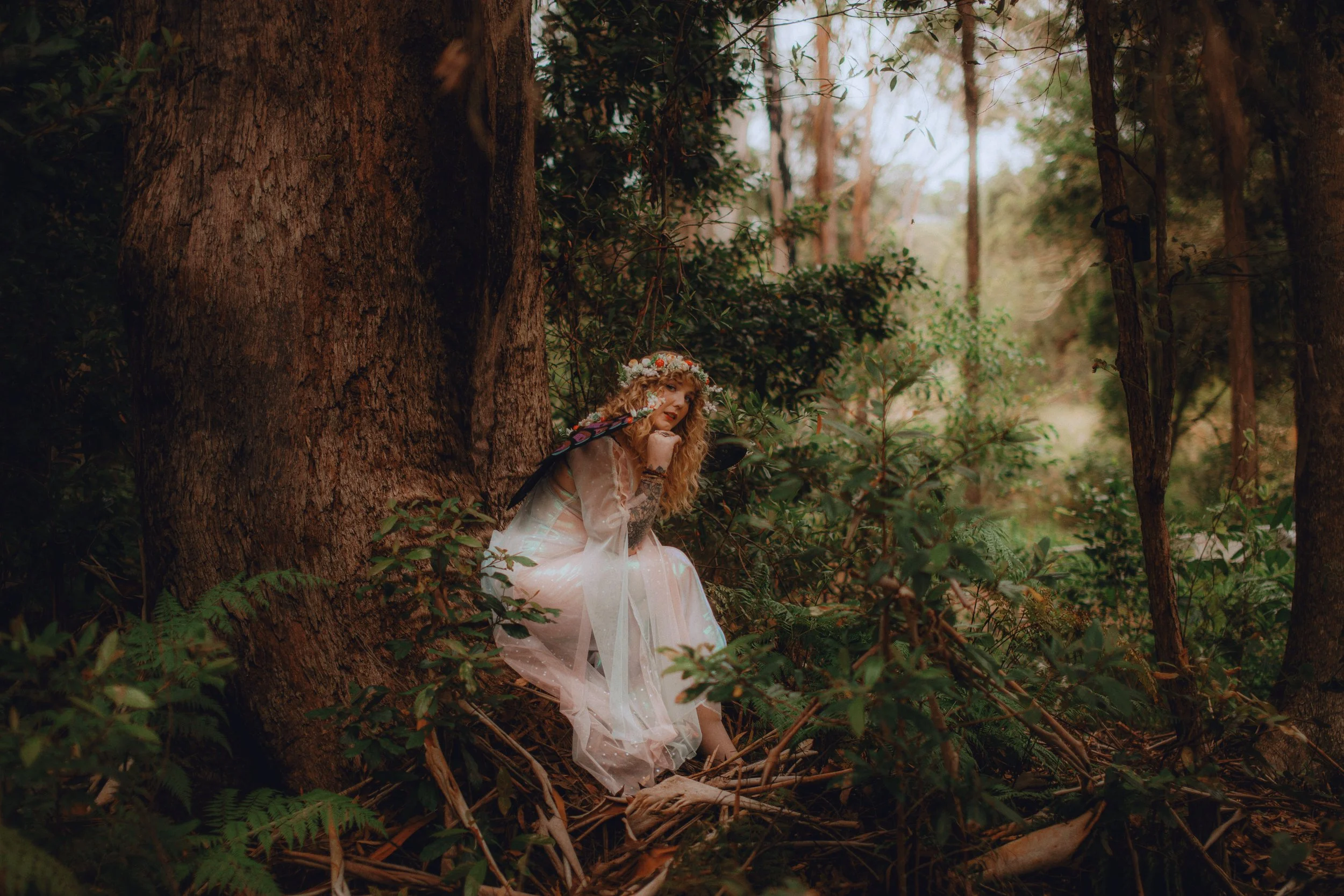 Woman dressed as a fairy, with a flower crown, sitting against a large tree trunk in a forest, surrounded by lush green foliage.