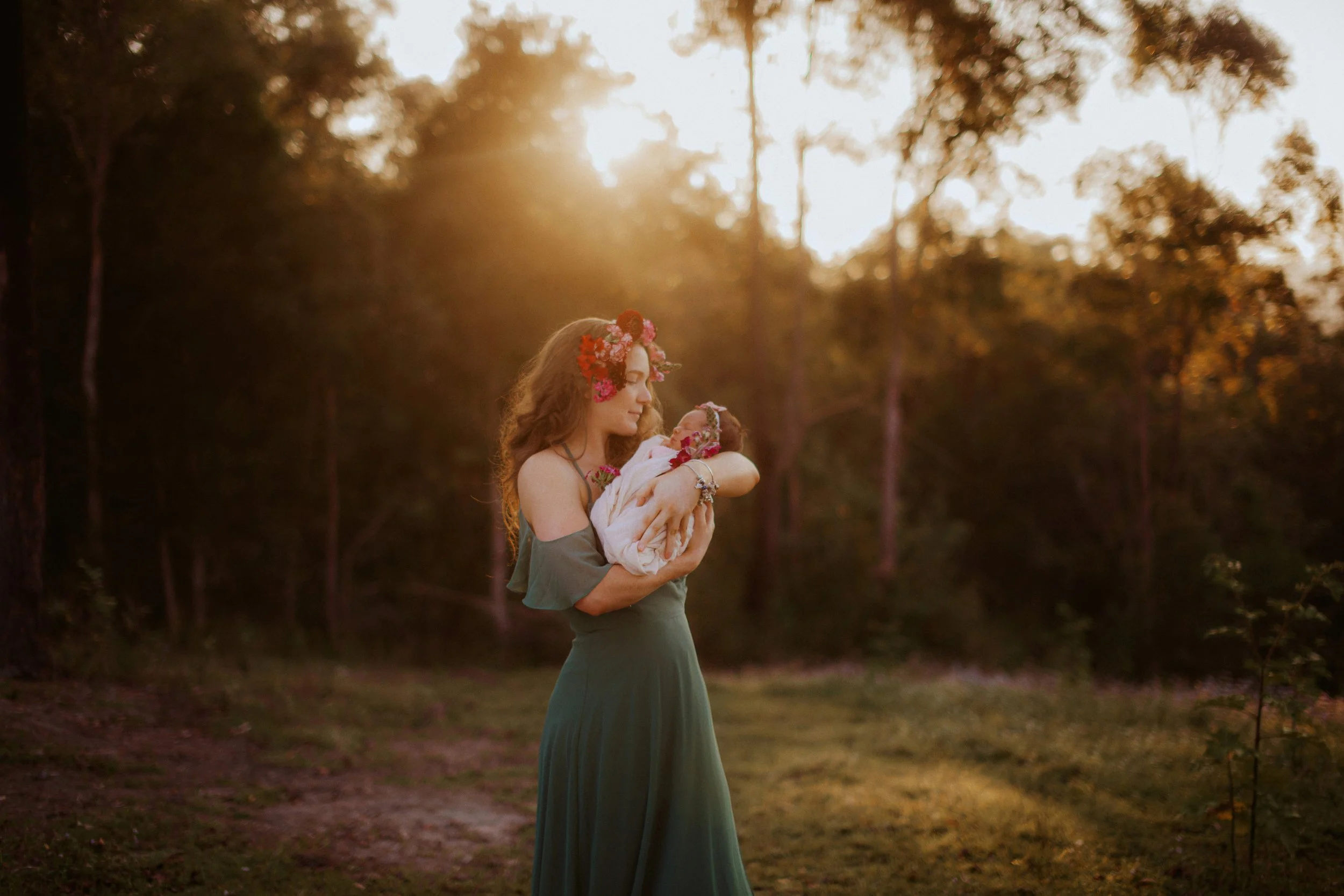 A woman in a long green dress holding a wrapped baby with flower decorations, standing outdoors during sunset in a wooded area.