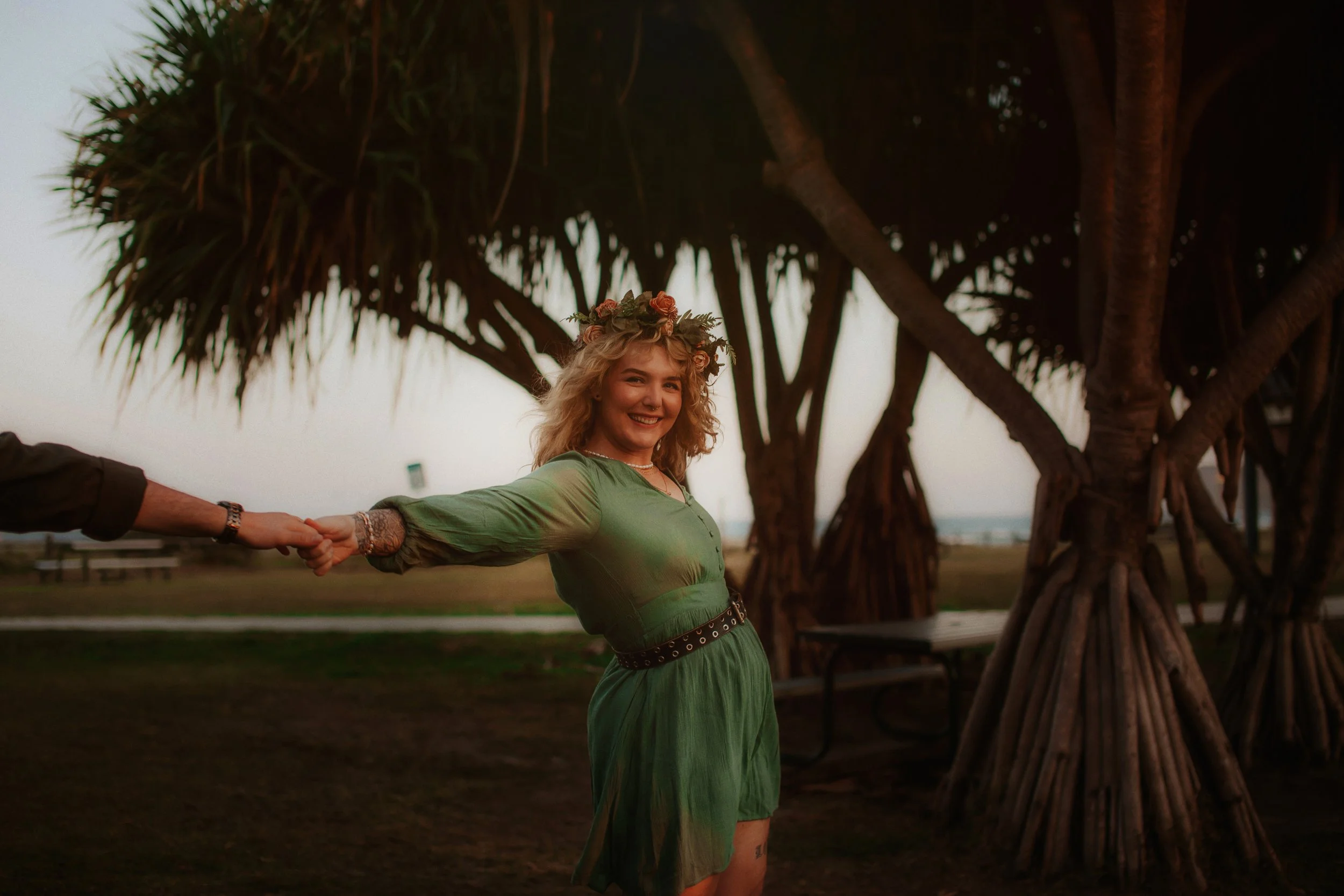A woman with curly blonde hair, wearing a green dress and a floral headband, holding hands with someone off-camera in an outdoor setting with large trees and a park bench in the background during sunset.
