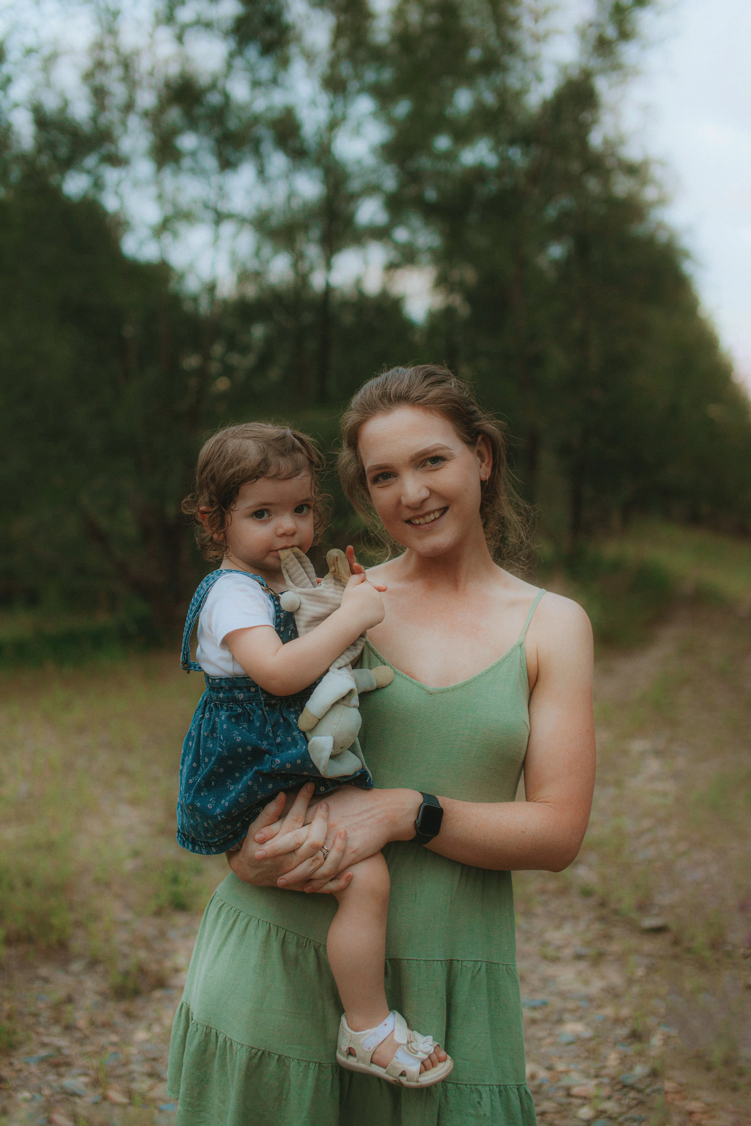 A woman with a young girl on her arm standing outdoors on a dirt path with trees in the background.