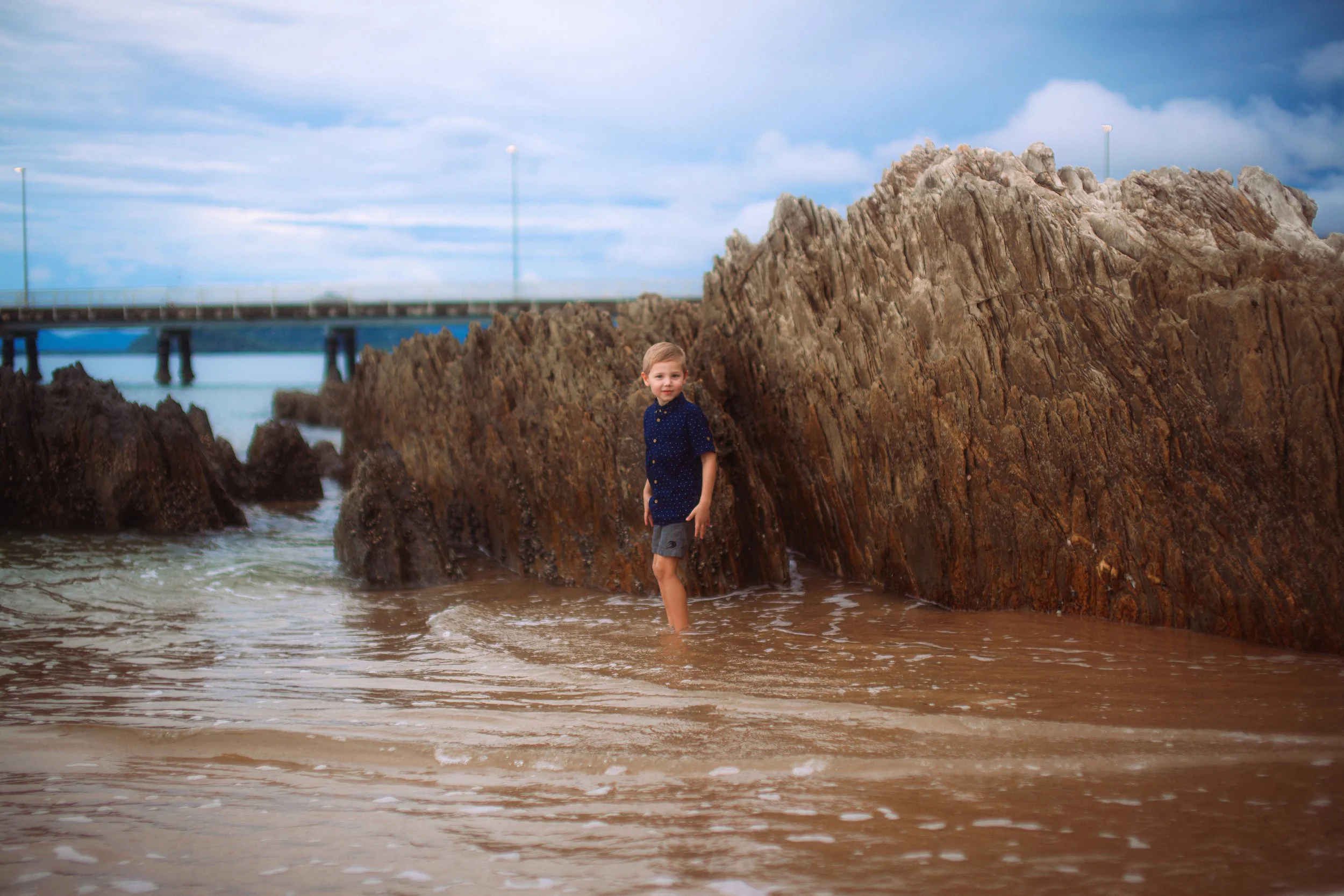 A young boy standing in shallow ocean water near a large rocky shoreline with a bridge in the background as cloudy skies overhead.