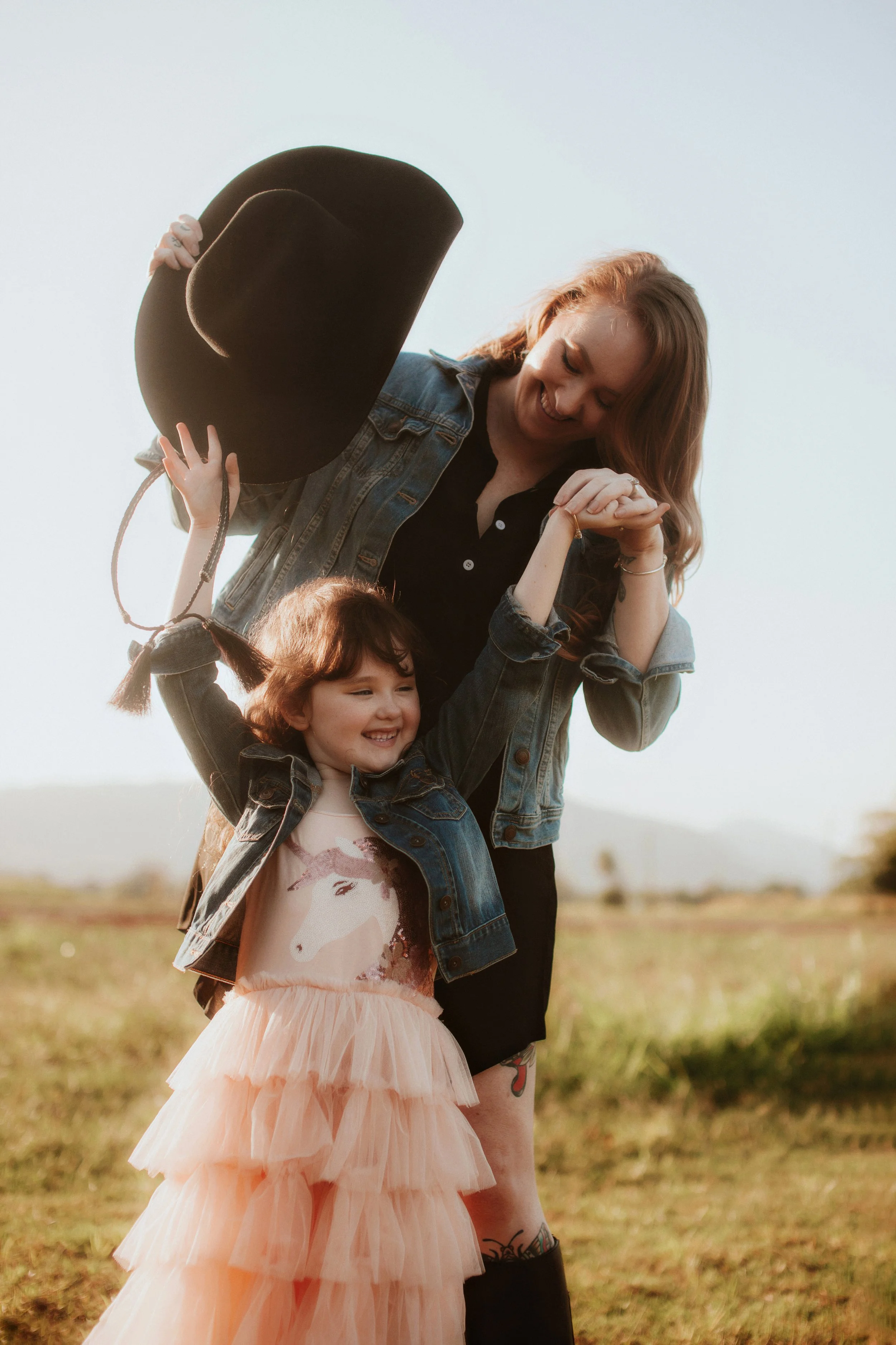 A woman and young girl enjoying a moment outdoors, both wearing denim jackets. The woman is holding a black wide-brimmed hat and the girl is wearing a peach-colored dress with a unicorn design.