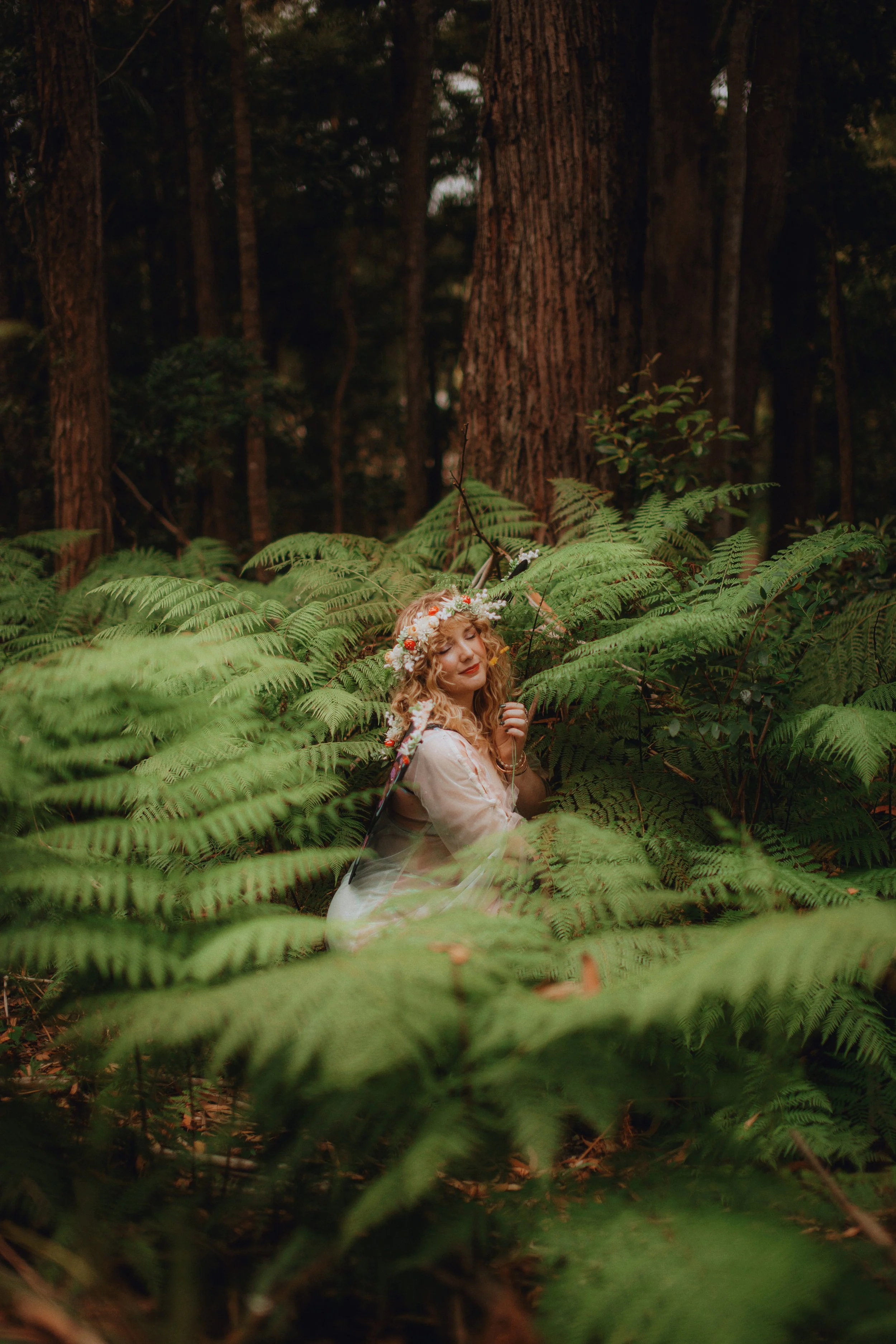 A woman with curly blonde hair, wearing a flower crown and a white dress, sitting among ferns in a forest.