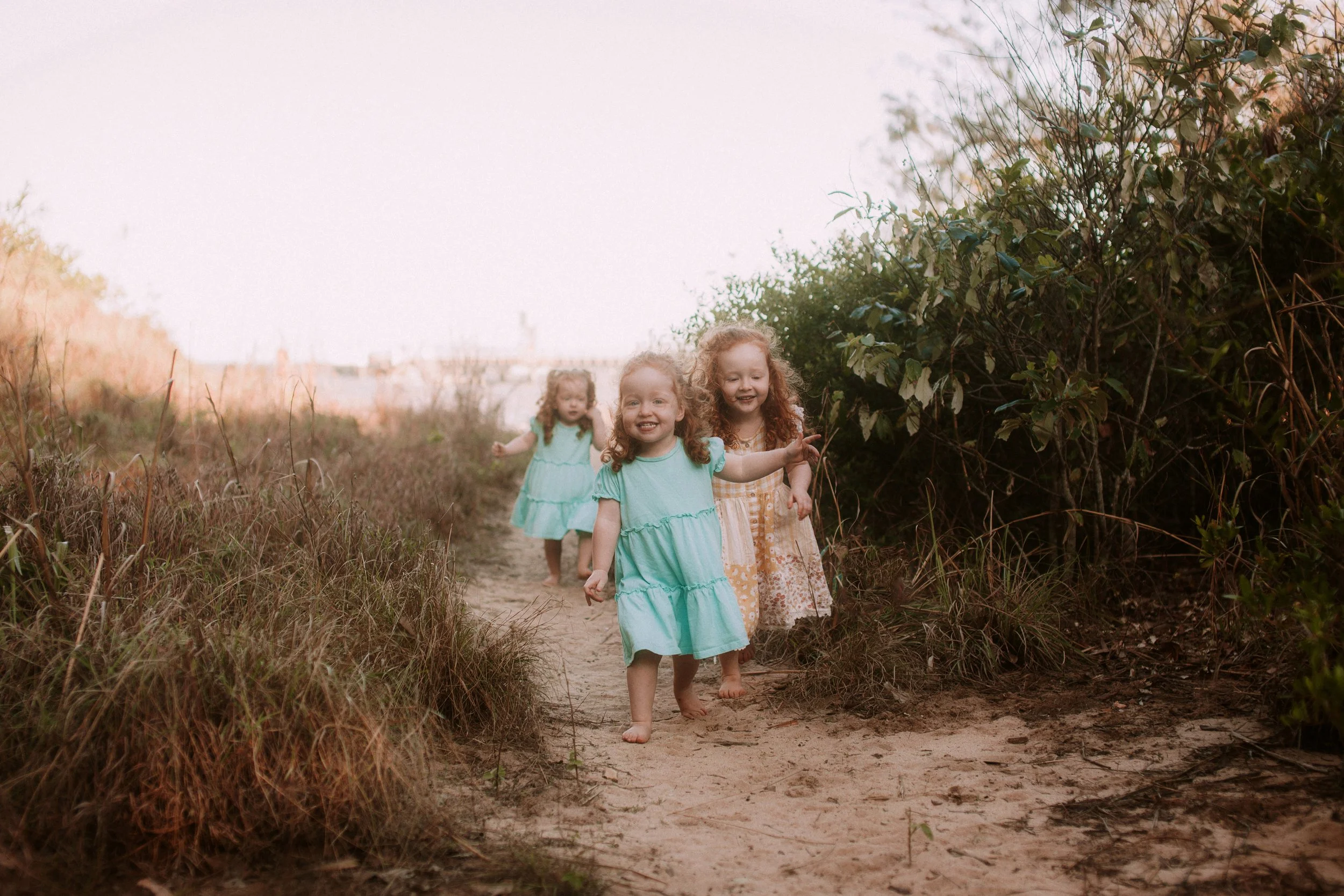 Three young girls in dresses walking barefoot on a sandy trail through a grassy area with bushes, smiling and enjoying outdoors in natural lighting.