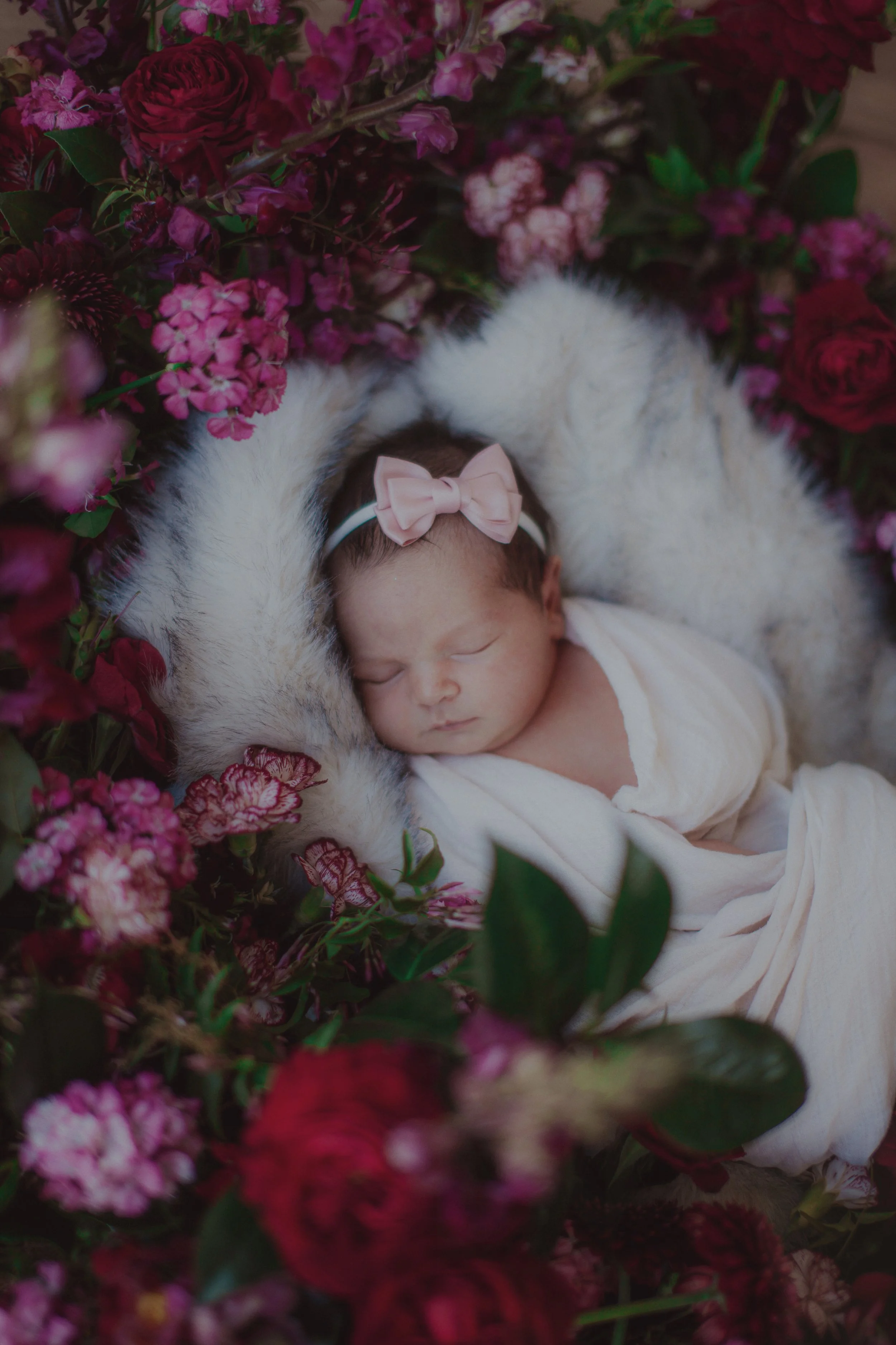 A newborn baby girl sleeping among pink, purple, and red flowers, wearing a white outfit and a pink bow headband.