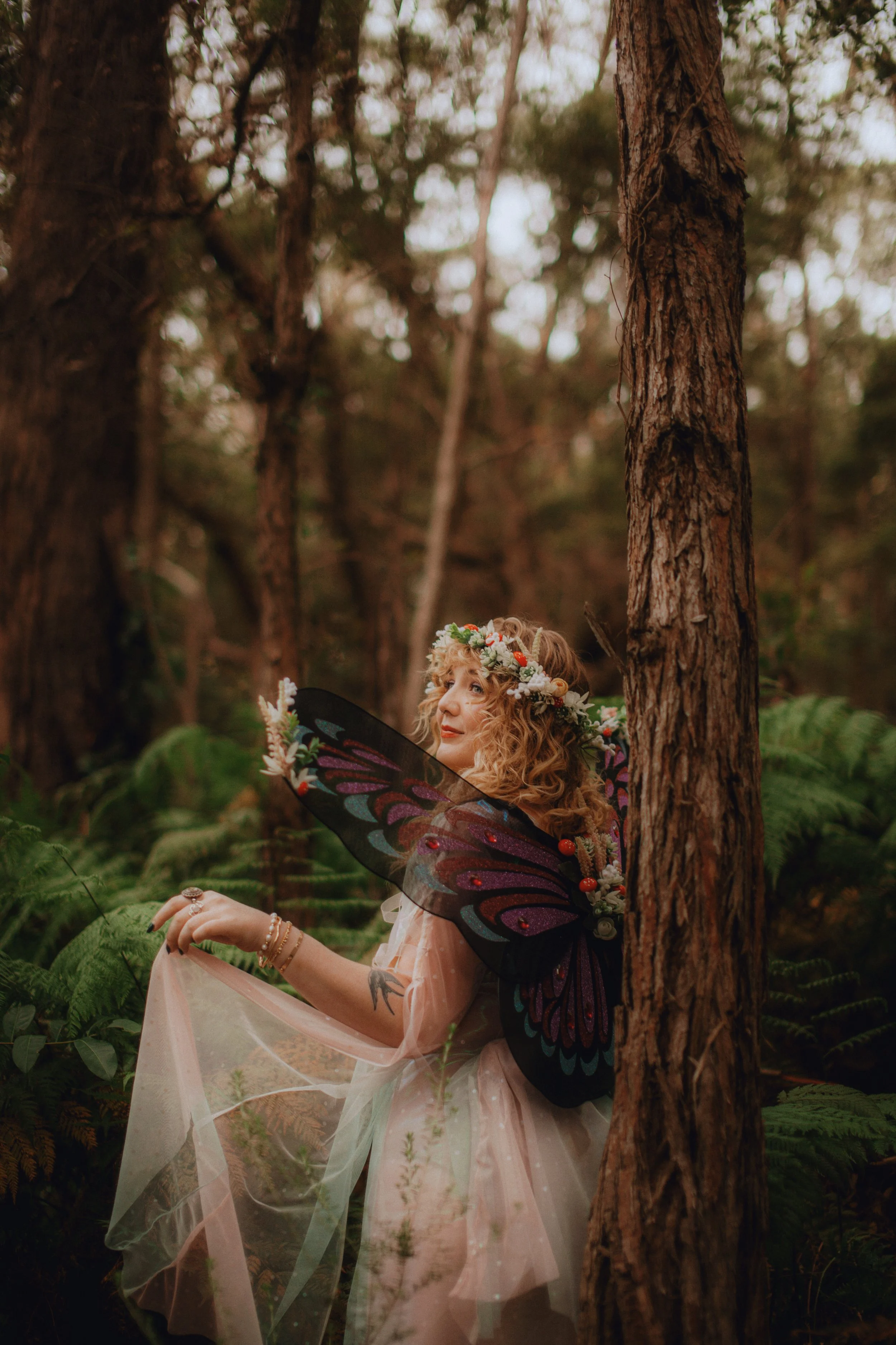 A woman with curly blonde hair wearing a floral crown and fairy wings stands in a lush forest surrounded by green ferns, holding a sheer pink fabric.