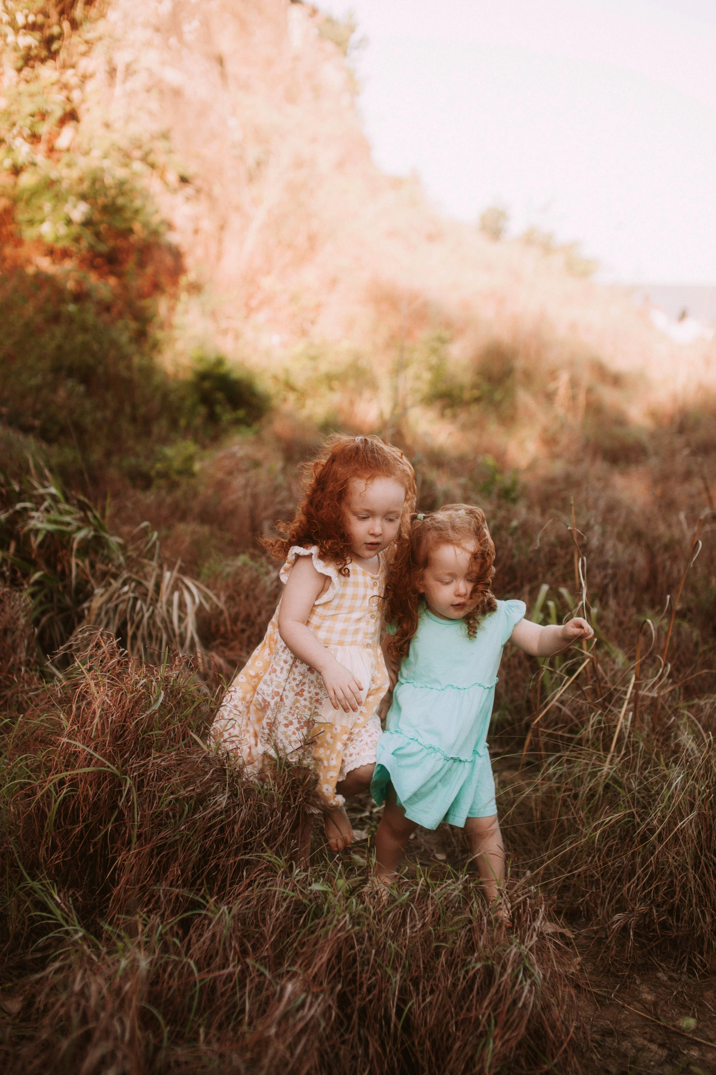Two young red-haired girls exploring a grassy outdoor area with tall grass and rocky cliffs in the background