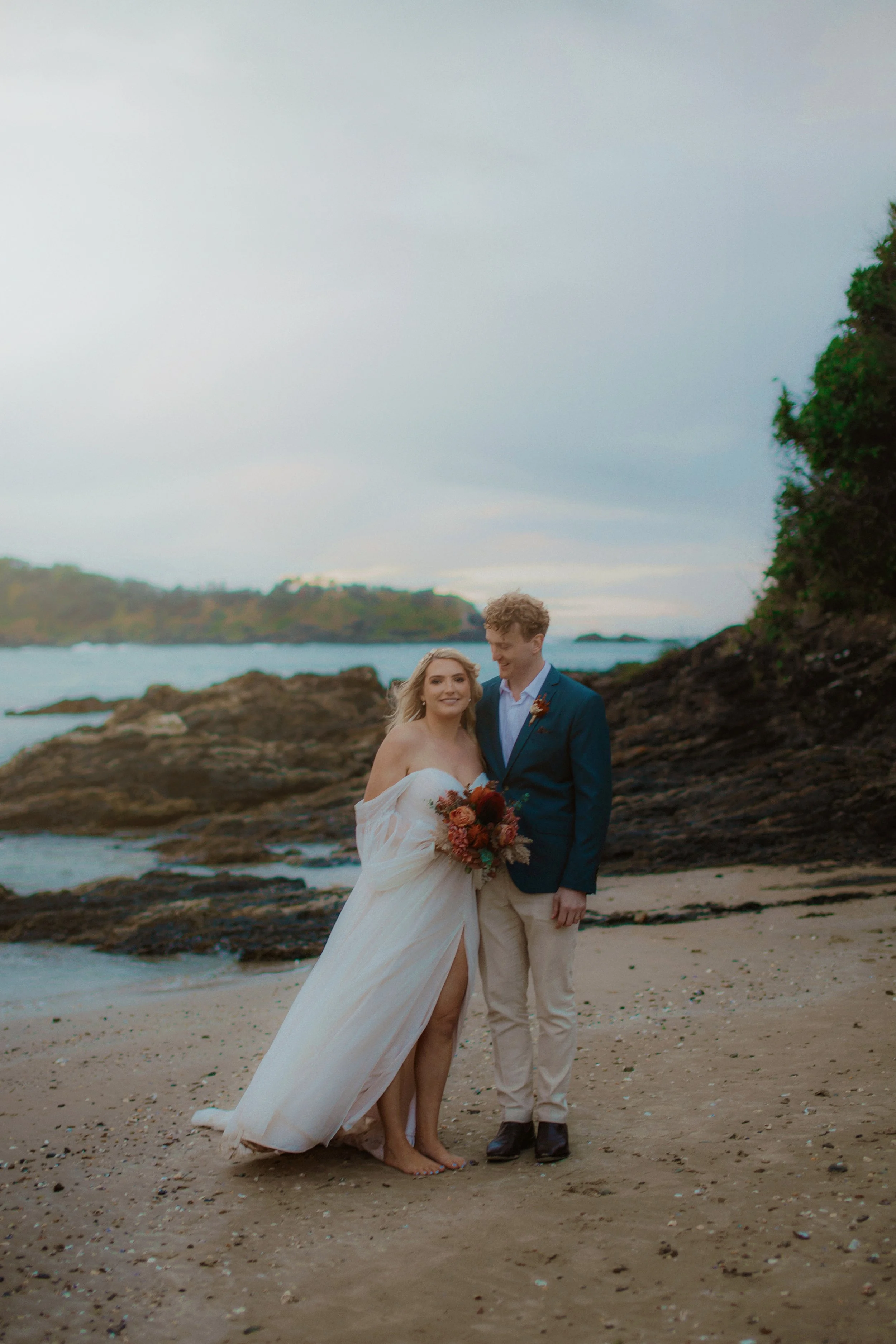 A bride and groom standing barefoot on a beach, smiling, with rocks and the ocean behind them.