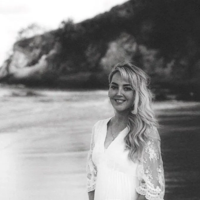 A woman with long wavy hair smiling at the camera, standing on a beach with rocky cliffs in the background.