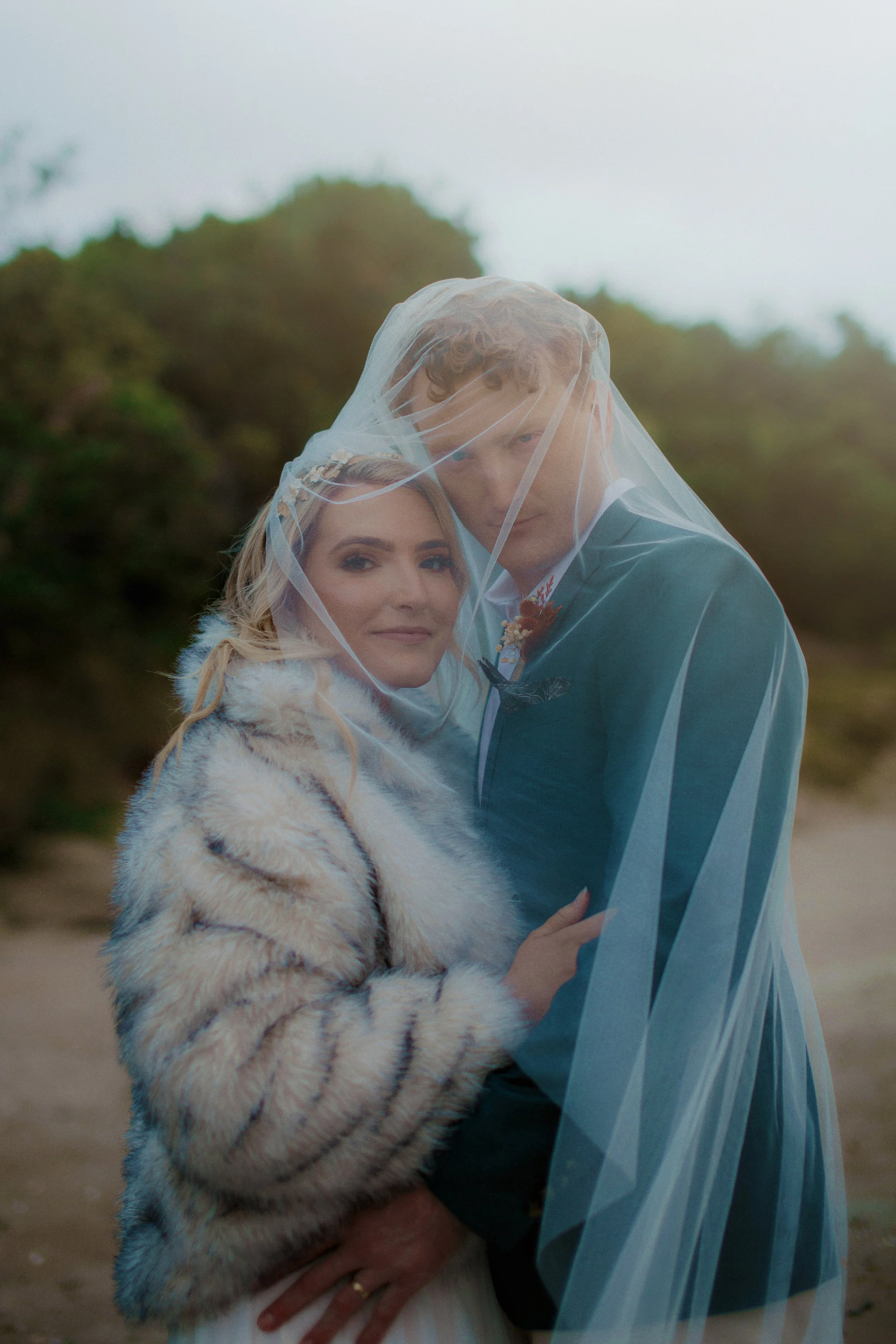 A bride and groom are close together on a beach, partially covered by a veil, with the bride wearing a fur coat and the groom in a blue suit.
