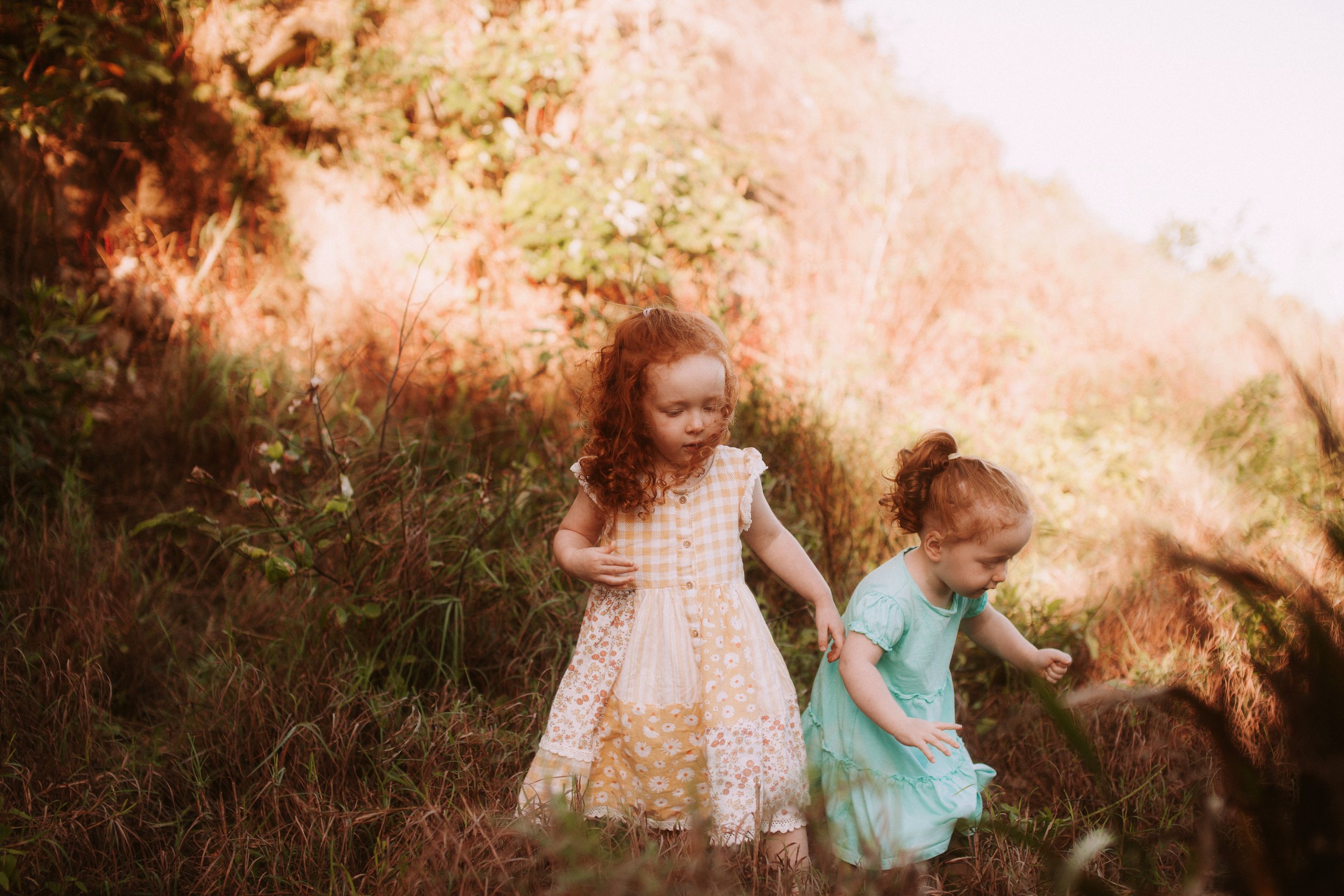 Two young red-haired girls, one in a yellow checkered dress and the other in a mint green dress, walking through a grassy field with trees in the background on a sunny day.