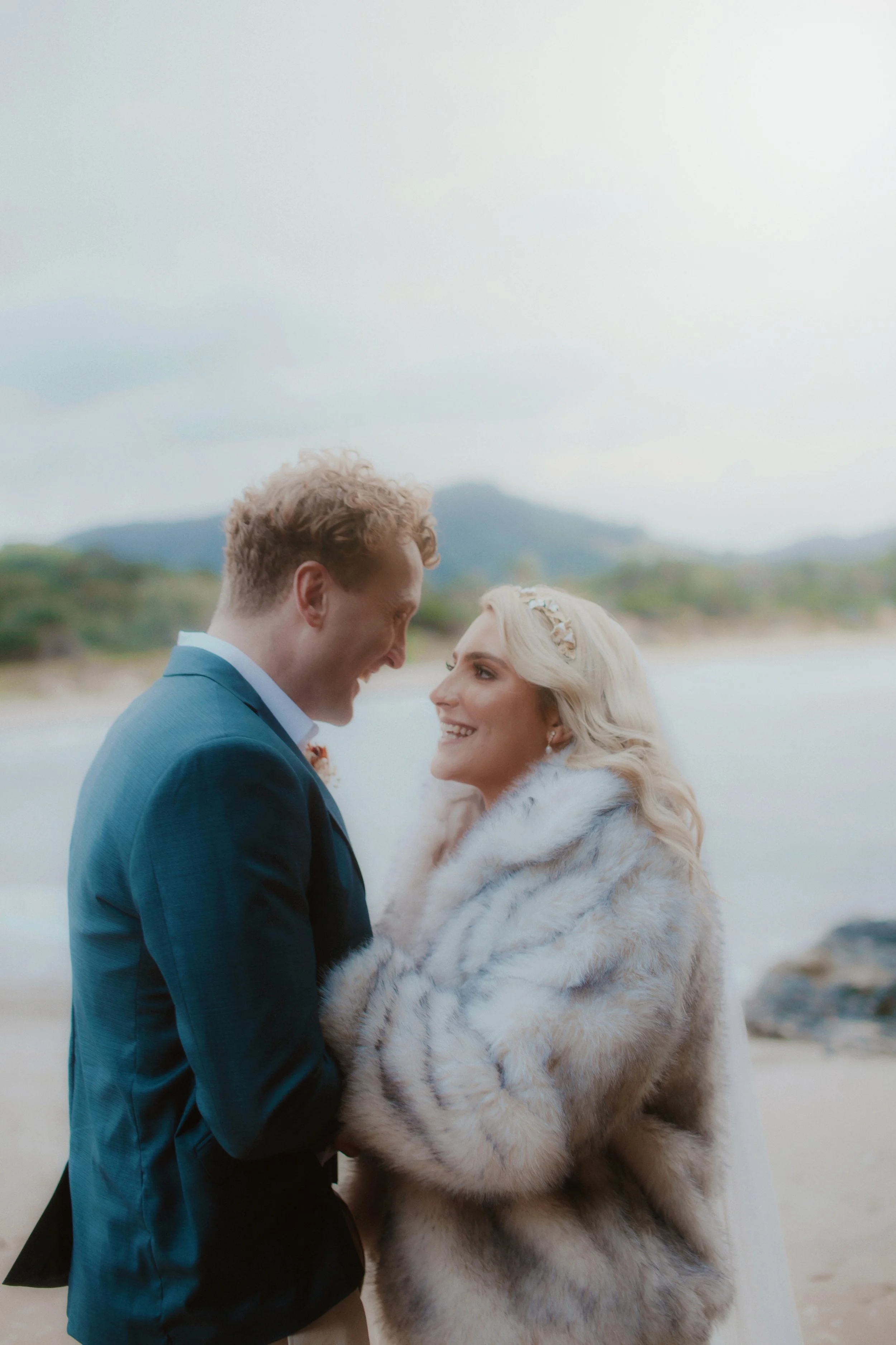 A bride and groom smiling at each other on the beach, with the groom wearing a blue suit and the bride in a fur coat, during their wedding.