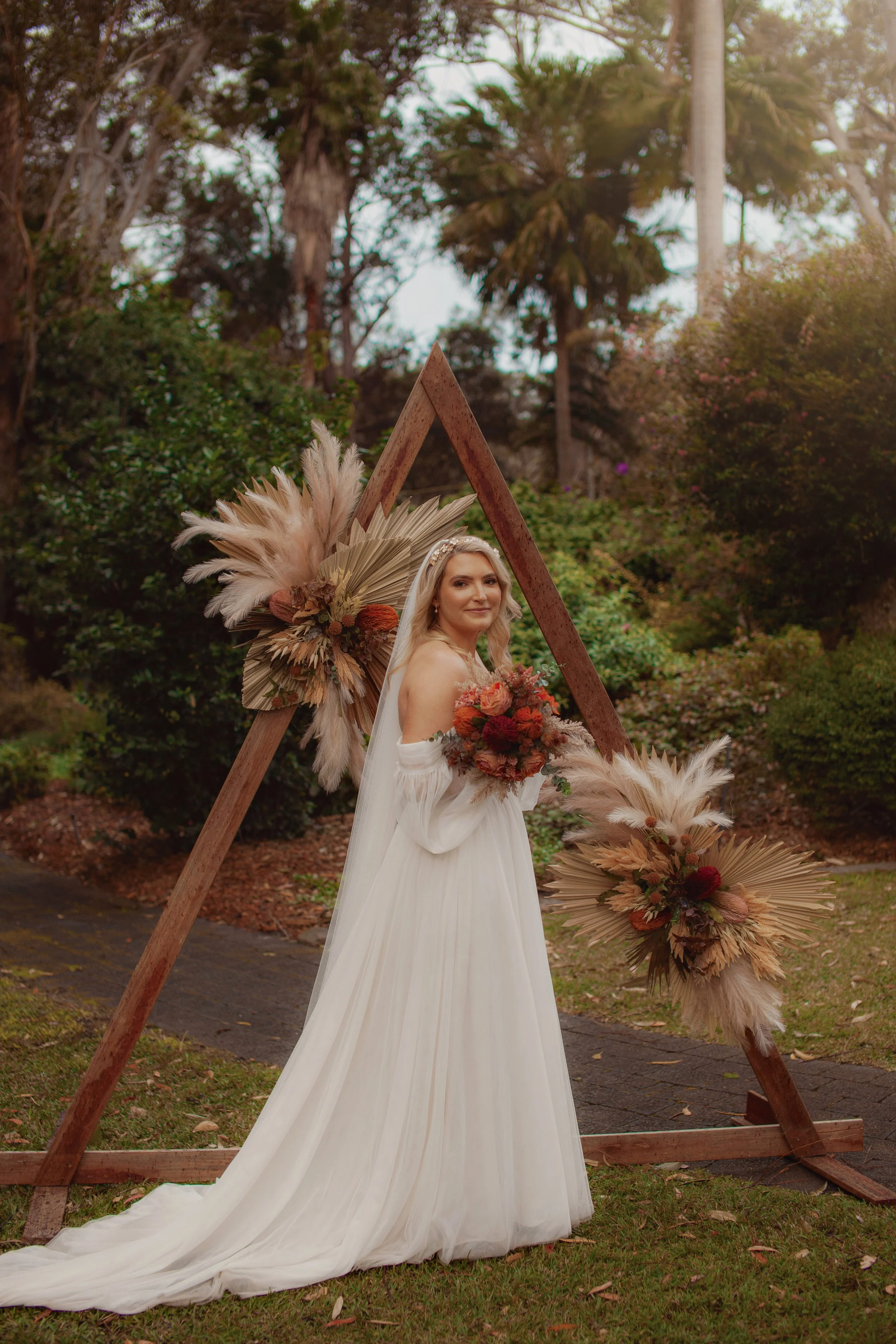Bride in a white gown holding a bouquet, standing next to a wooden wedding arch decorated with dried flowers and pampas grass, outdoors among trees and greenery.