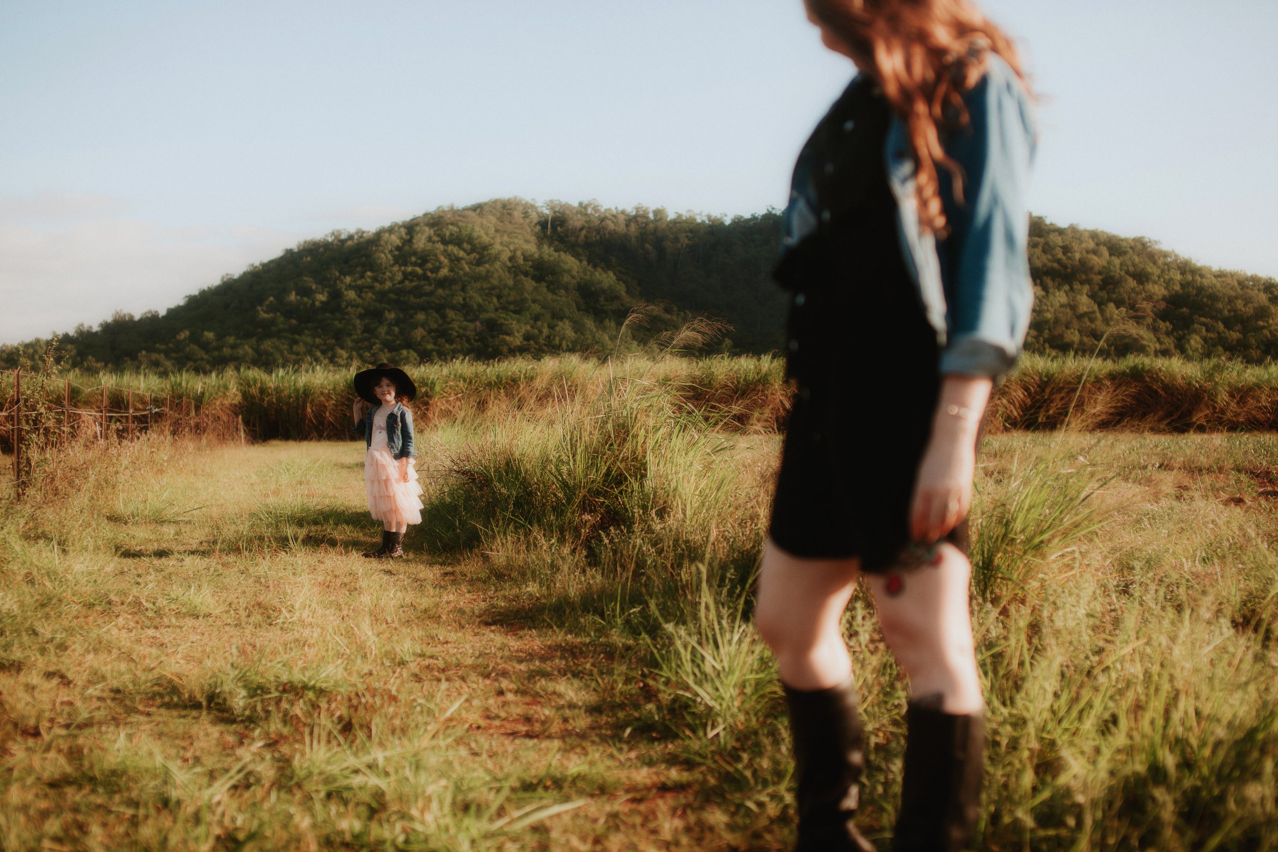Two women outdoors, one in the foreground with knee-high boots and colorful clothing, and one in the background wearing a large black hat, light-colored dress, and denim jacket, standing on a grassy path with a mountain in the distance.
