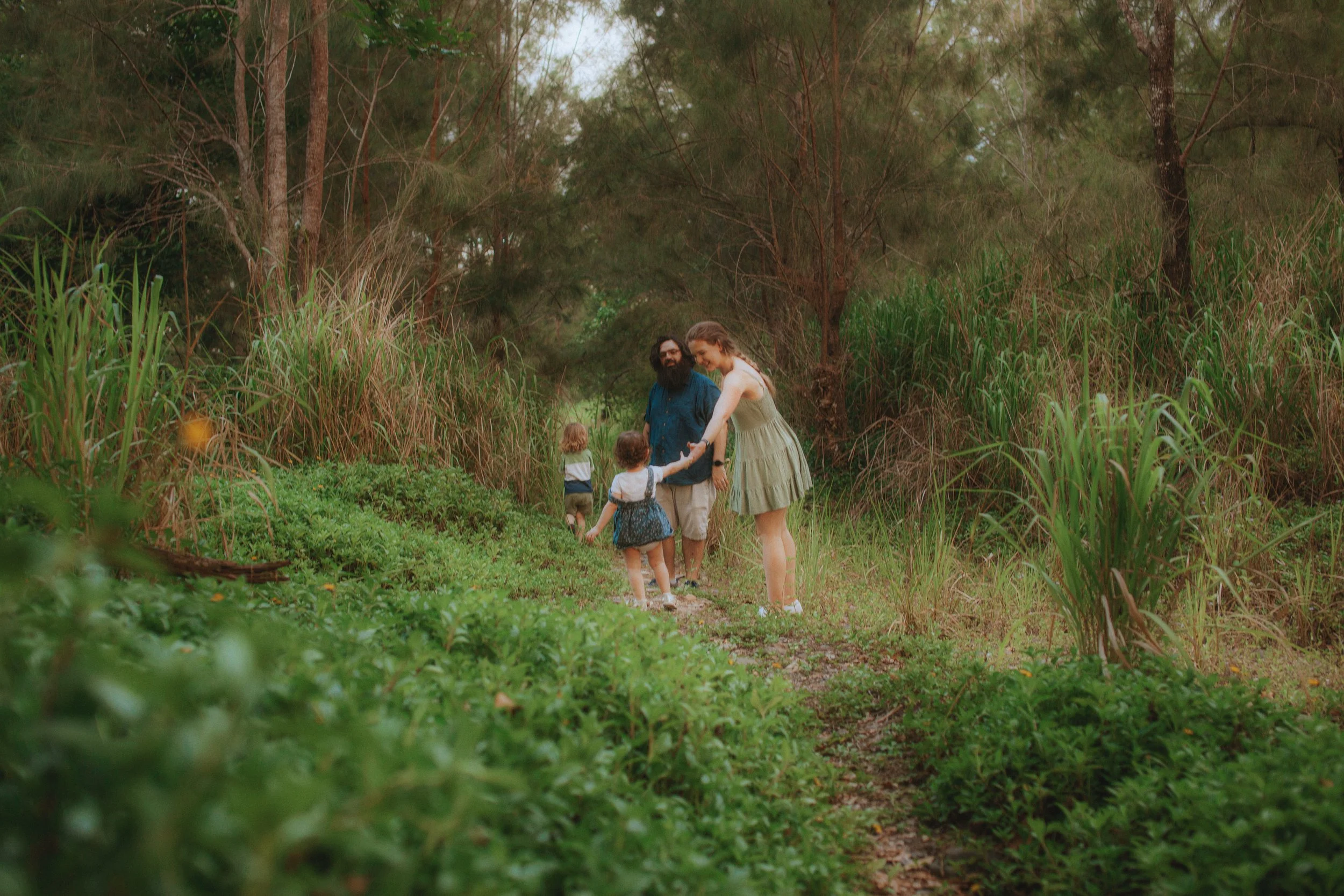 A family of four, including a woman, a man, and two young children, walking along a narrow dirt path through a lush green forest with tall grass and trees.