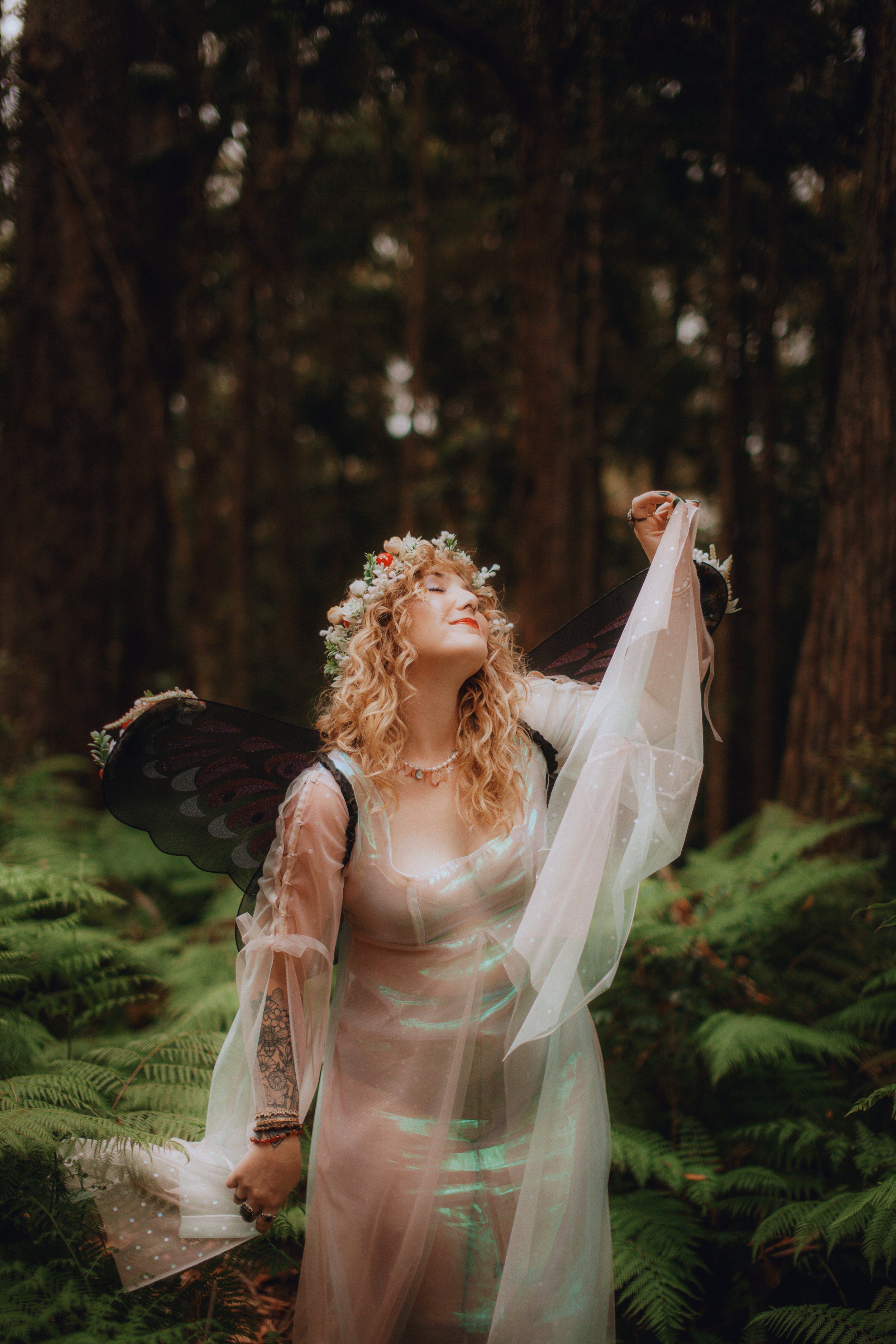A woman dressed as a fairy with black butterfly wings, a floral crown, and a flowing gown, standing in a lush forest with ferns, holding a sheer fabric and looking content.