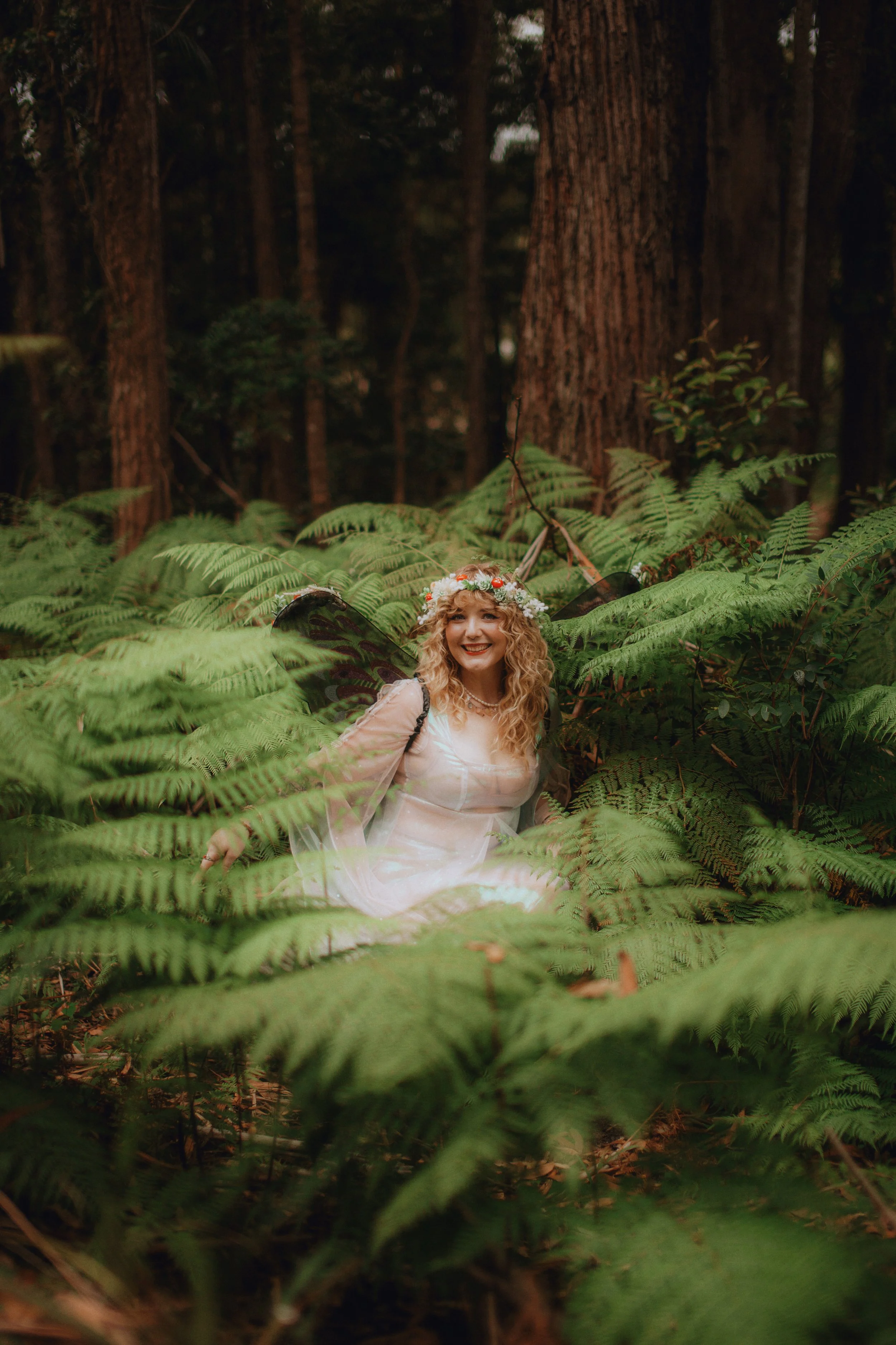 A woman dressed as a fairy with butterfly wings and a flower crown sitting among lush green ferns in a forest, smiling happily.