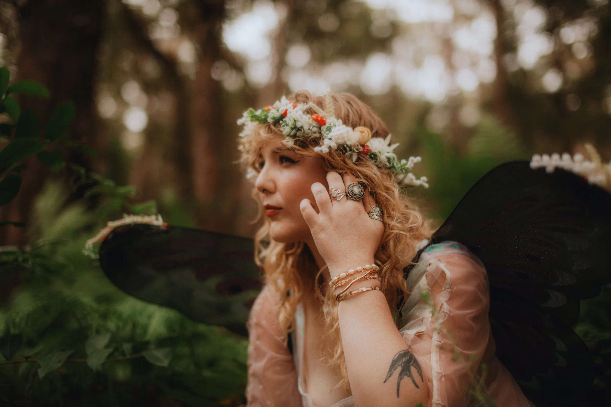 A woman with curly blonde hair wearing a flower crown and black fairy wings, touching her face with her left hand. She has rings, bracelets, and a tattoo on her left arm, and is in a forest with trees and greenery.