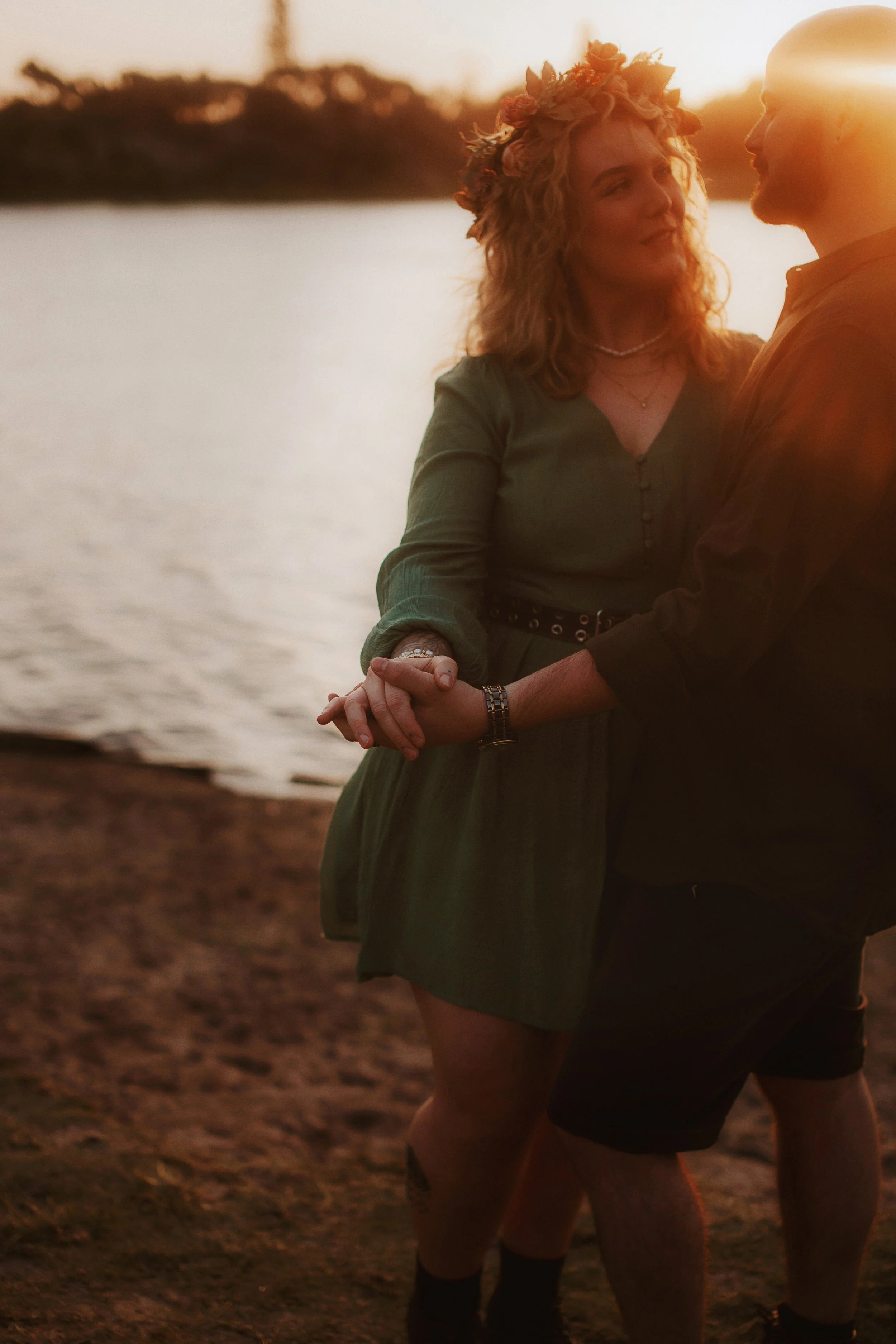 A couple holding hands by a lake at sunset, with the woman wearing a green dress and a flower crown, and the man wearing a dark shirt and shorts.