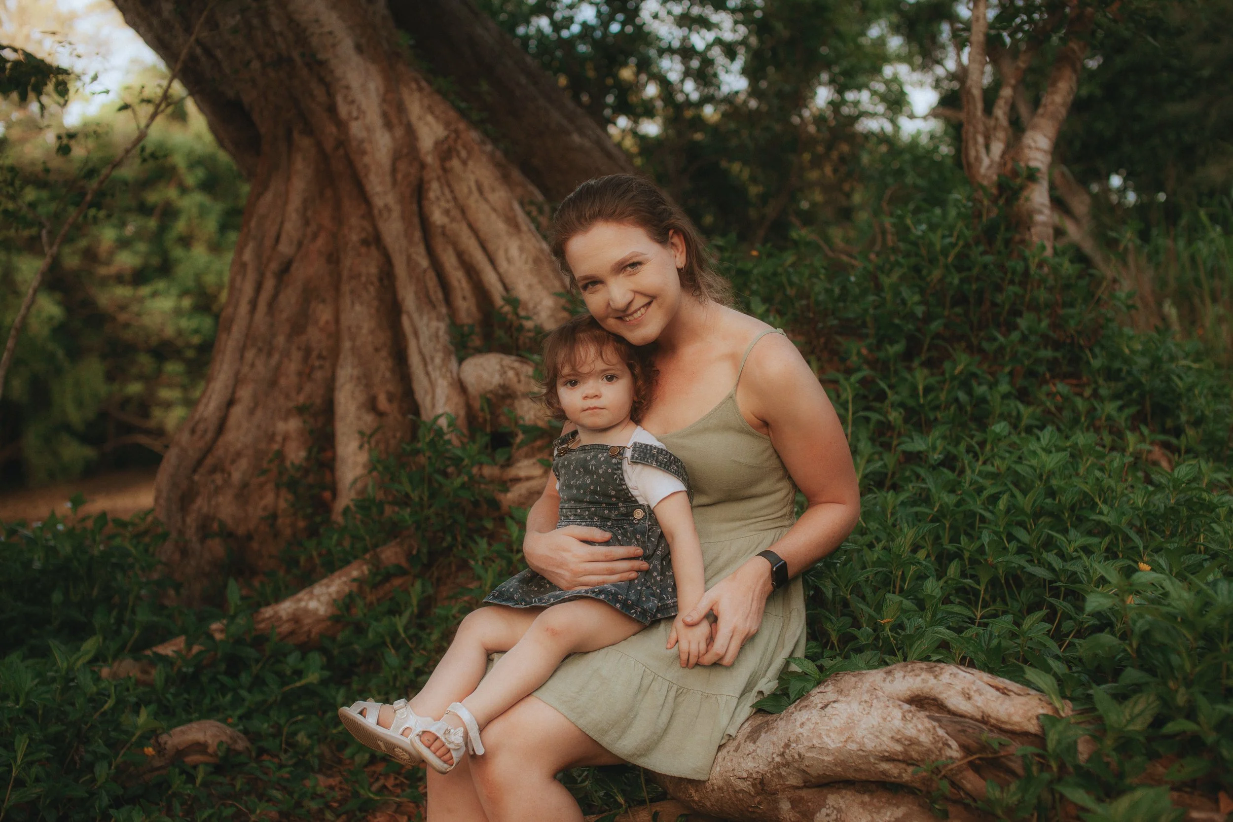A smiling woman sitting on a tree root holding a young girl on her lap in a lush green forest setting.