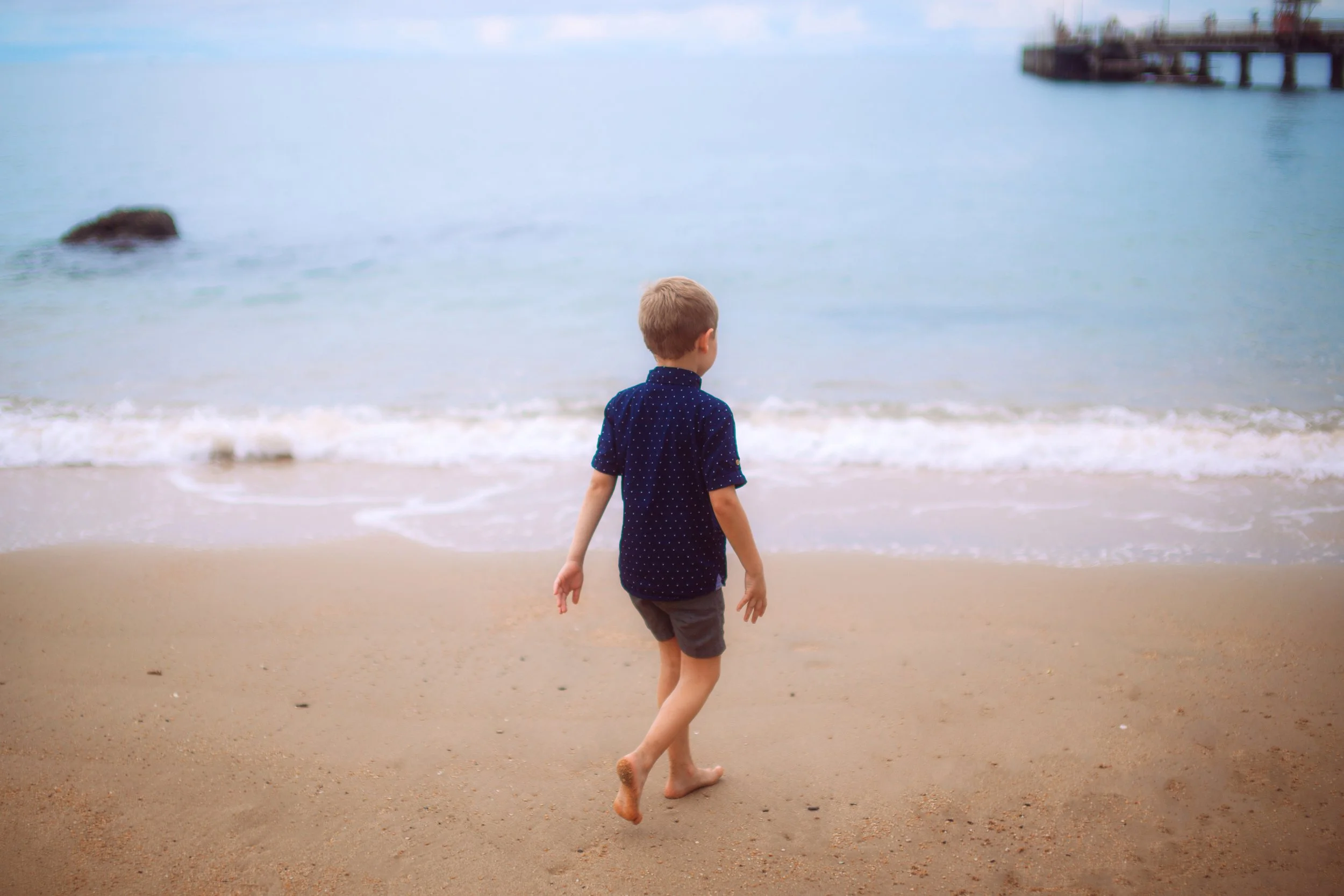 A young boy walking barefoot on the sandy beach near the ocean, with a pier in the distance.